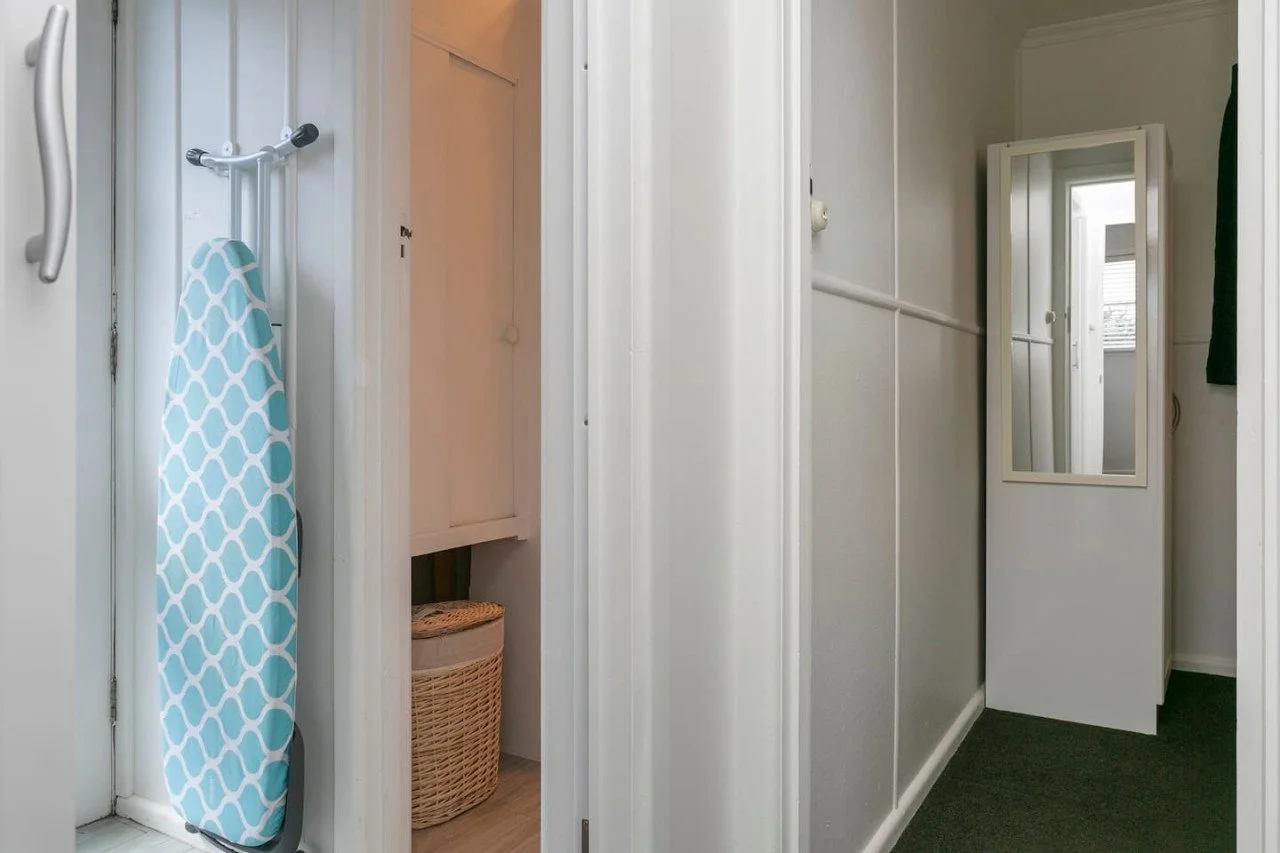 Interior view of a hallway with a full-length mirror and a small storage cabinet. An ironing board with a blue and white cover leans against the wall near a closet.