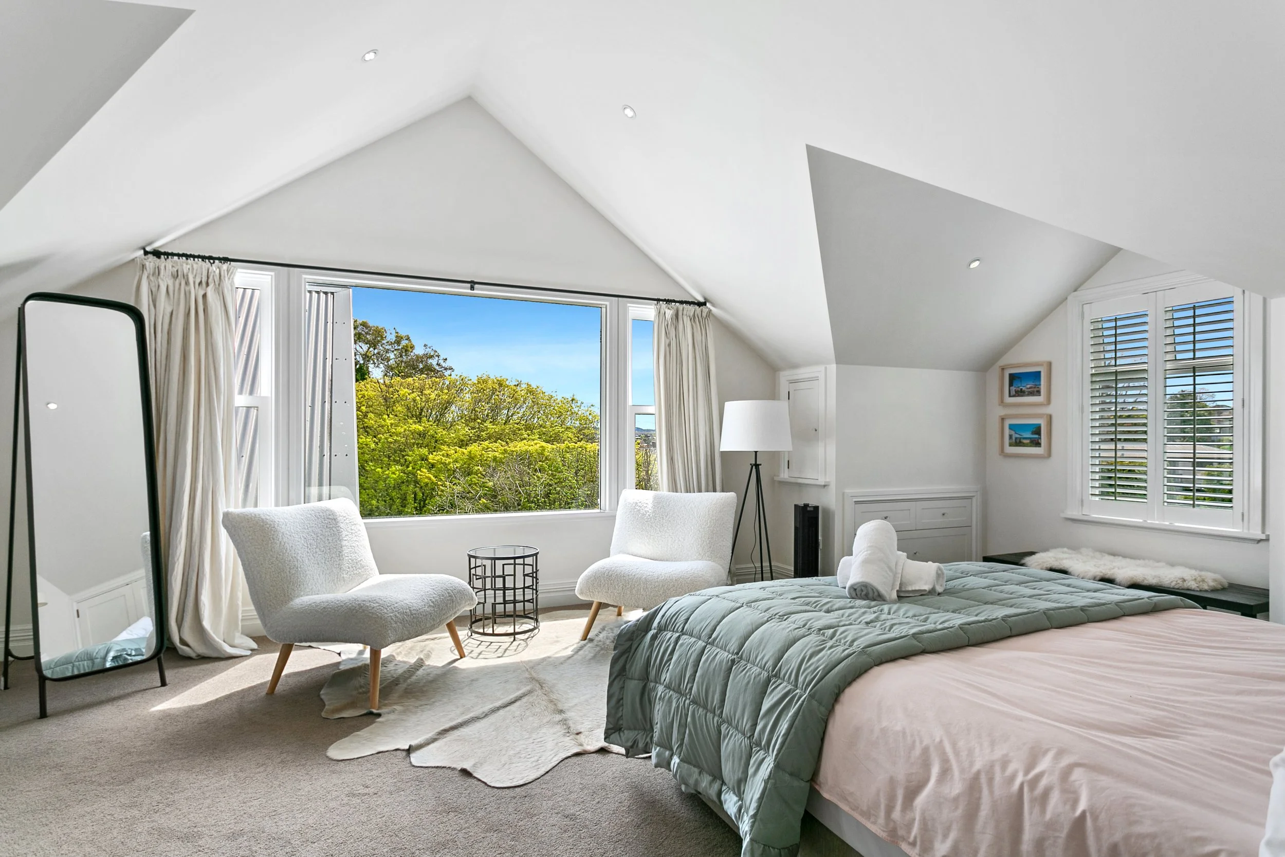 Bright bedroom with large window showing green trees, two white chairs, a round side table, a floor lamp, a bed with rolled towels, and window shutters.