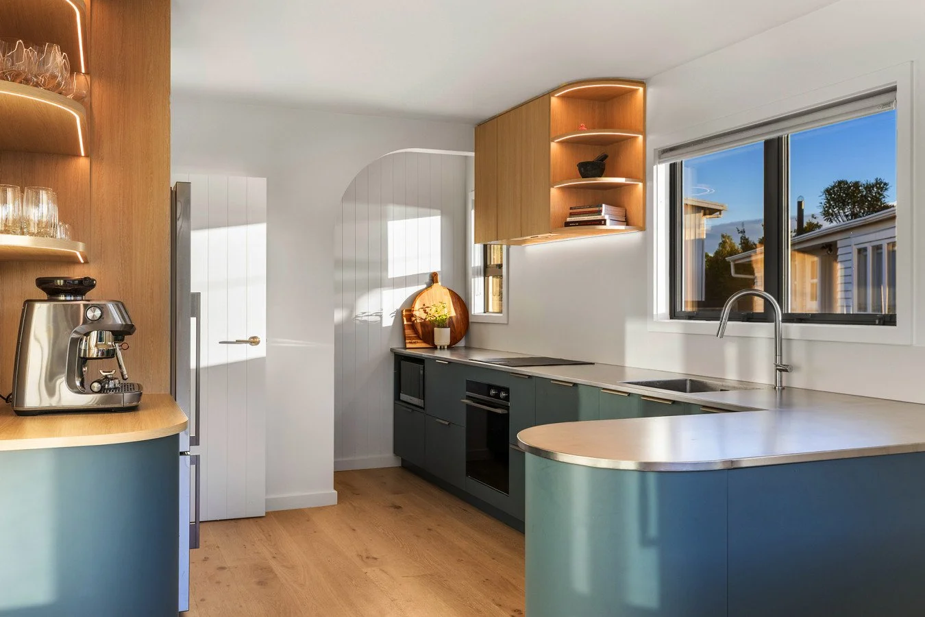 Modern kitchen with blue cabinets, wooden shelves, a coffee machine, and a large window showing a sunset sky.