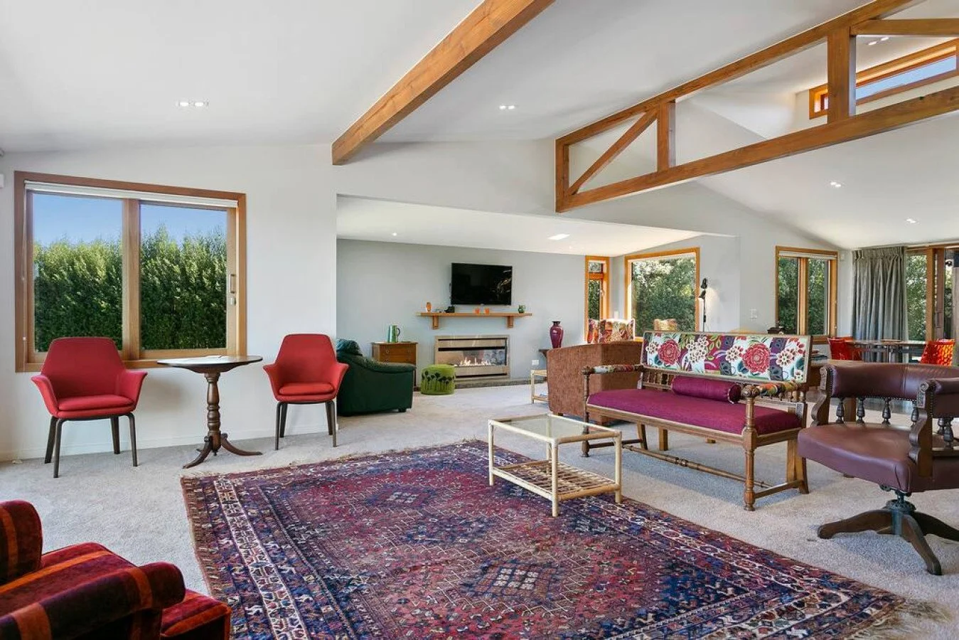Spacious living room with large windows, wooden beams, colorful furniture, and a patterned rug.