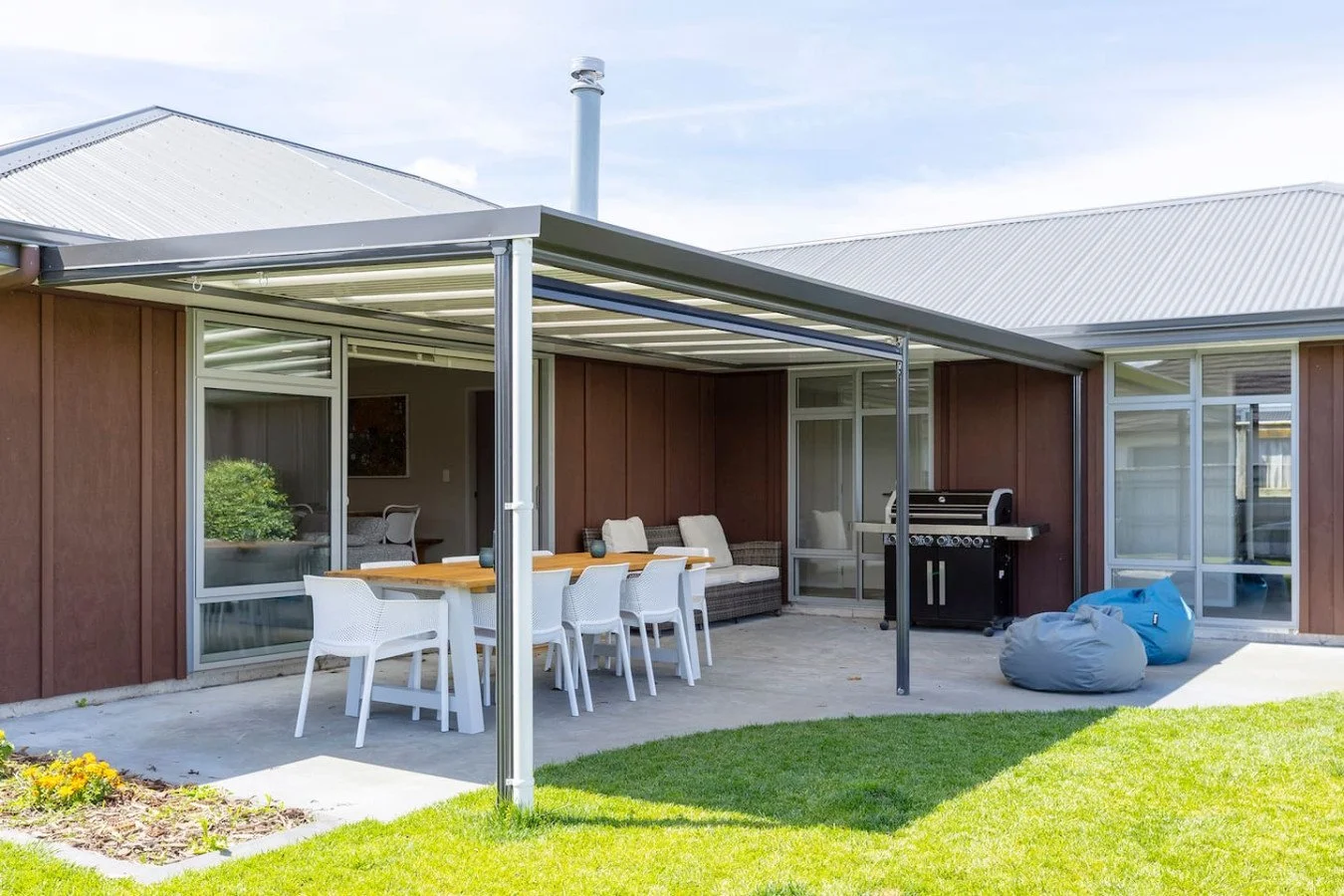 Backyard patio with outdoor dining table, chairs, lounge seating, propane grill, and bean bags, covered by a metal pergola with a house in the background.