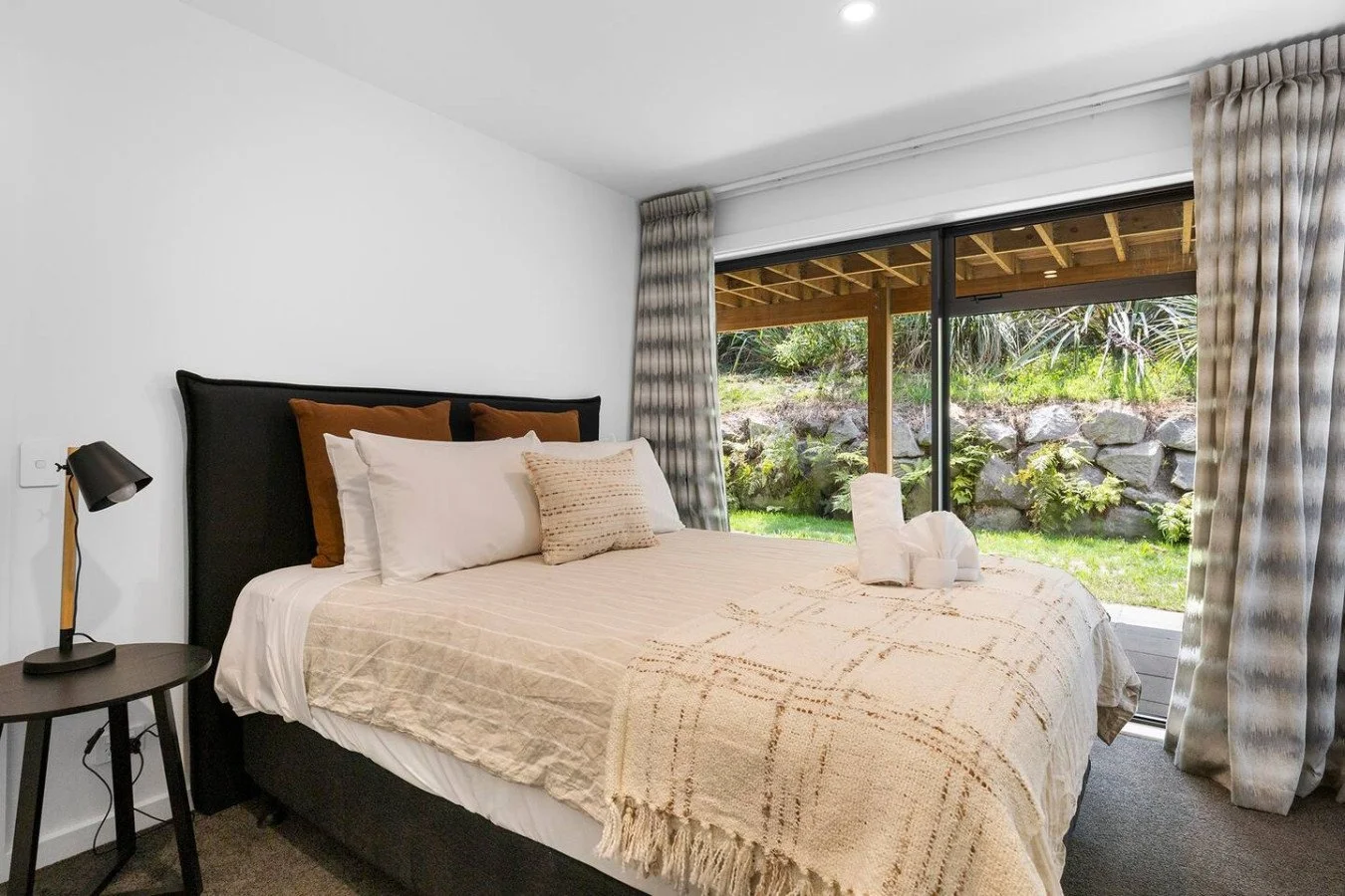 A neatly made bed with pillows and a beige throw blanket in a modern bedroom with sliding glass doors opening to a garden with rocks and plants.