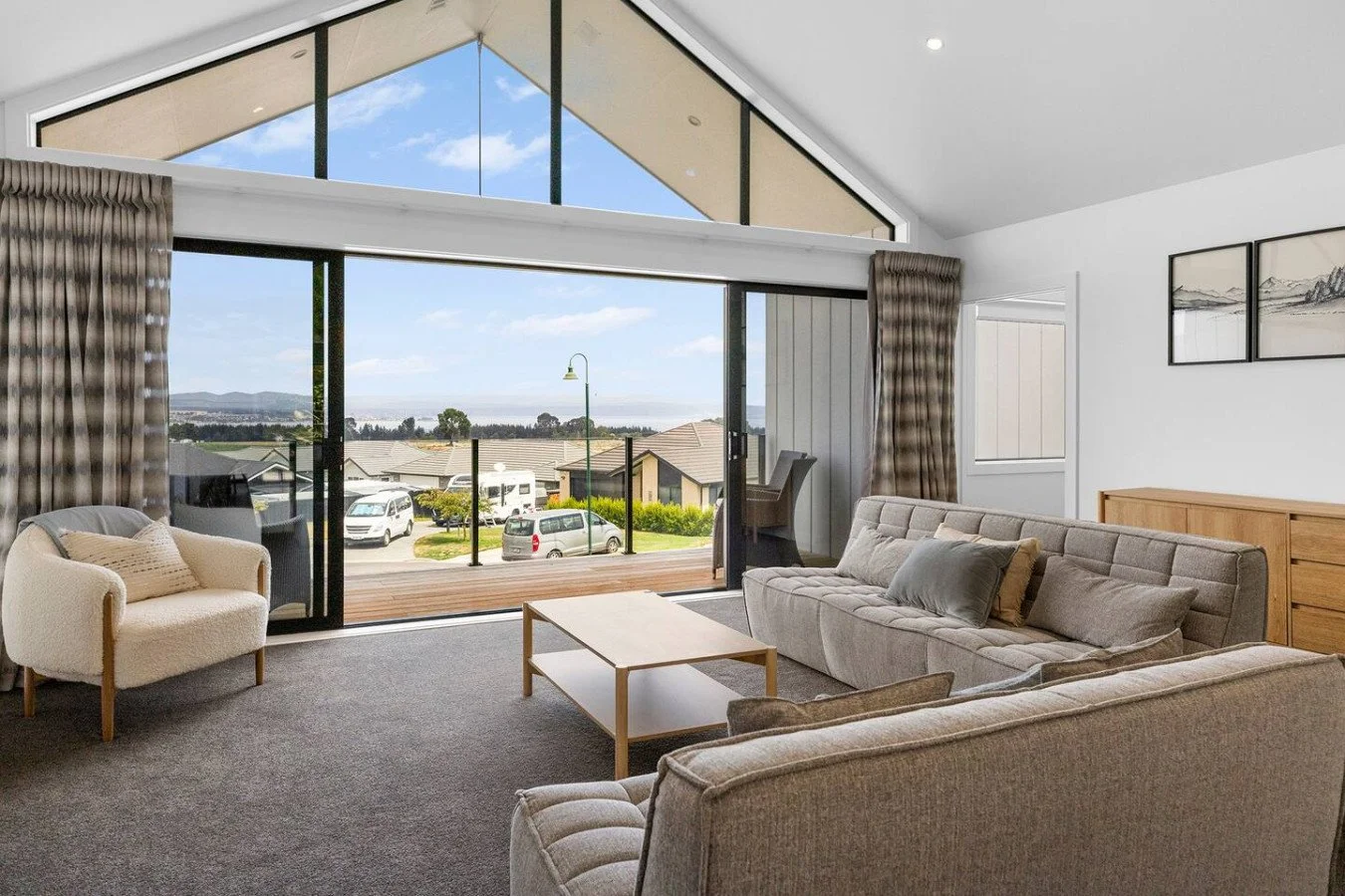 Living room with large glass sliding doors leading to a balcony, view of parked cars and houses outside, and mountains in the distance, furnished with beige sofas, an armchair, a coffee table, and curtains.