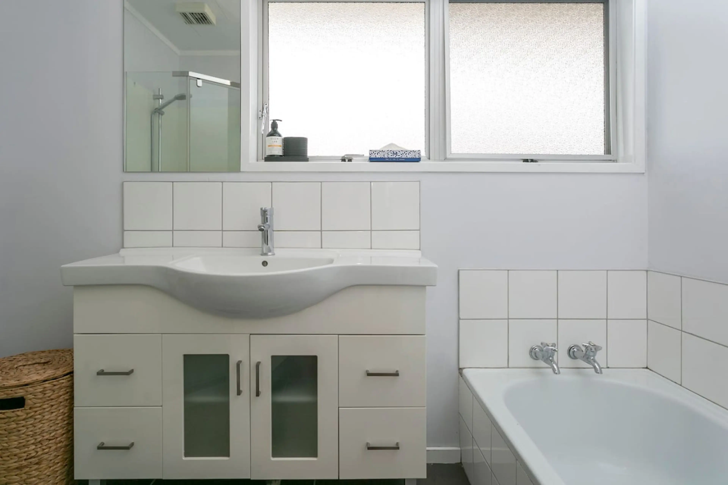 A modern bathroom with a white sink cabinet, a white tub with wall-mounted faucets, a mirror, and a frosted window with a soap dispenser and black containers on the windowsill.
