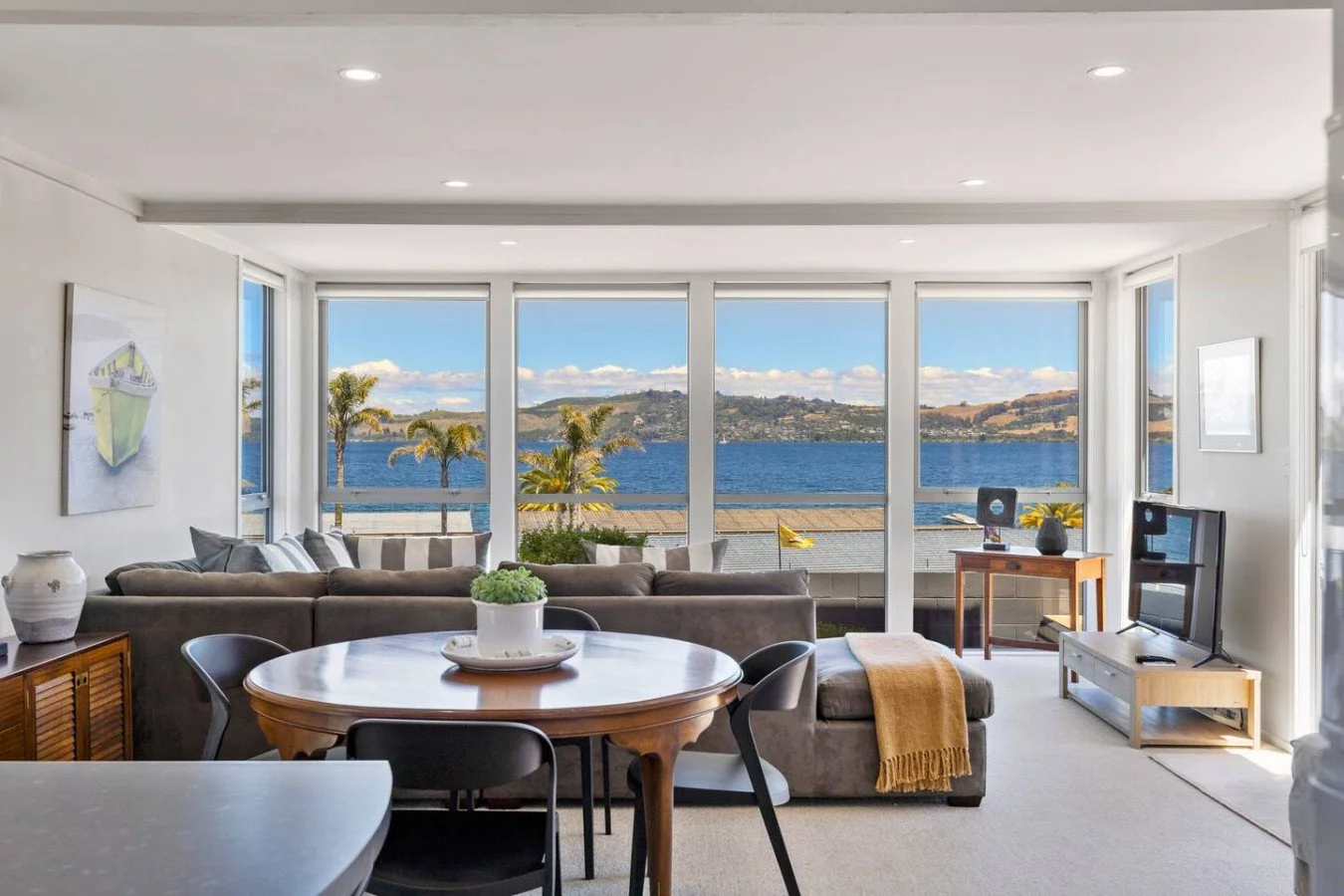 Living room with large windows overlooking water, with palm trees, sofa, dining table, and TV.