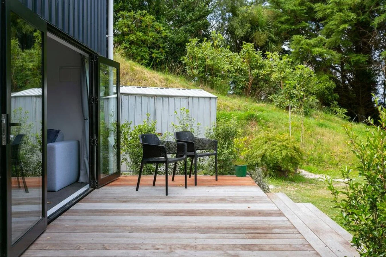 View of a wooden patio with two black chairs, a sliding glass door to a living room, and lush green trees and shrubs on a hill in the background.