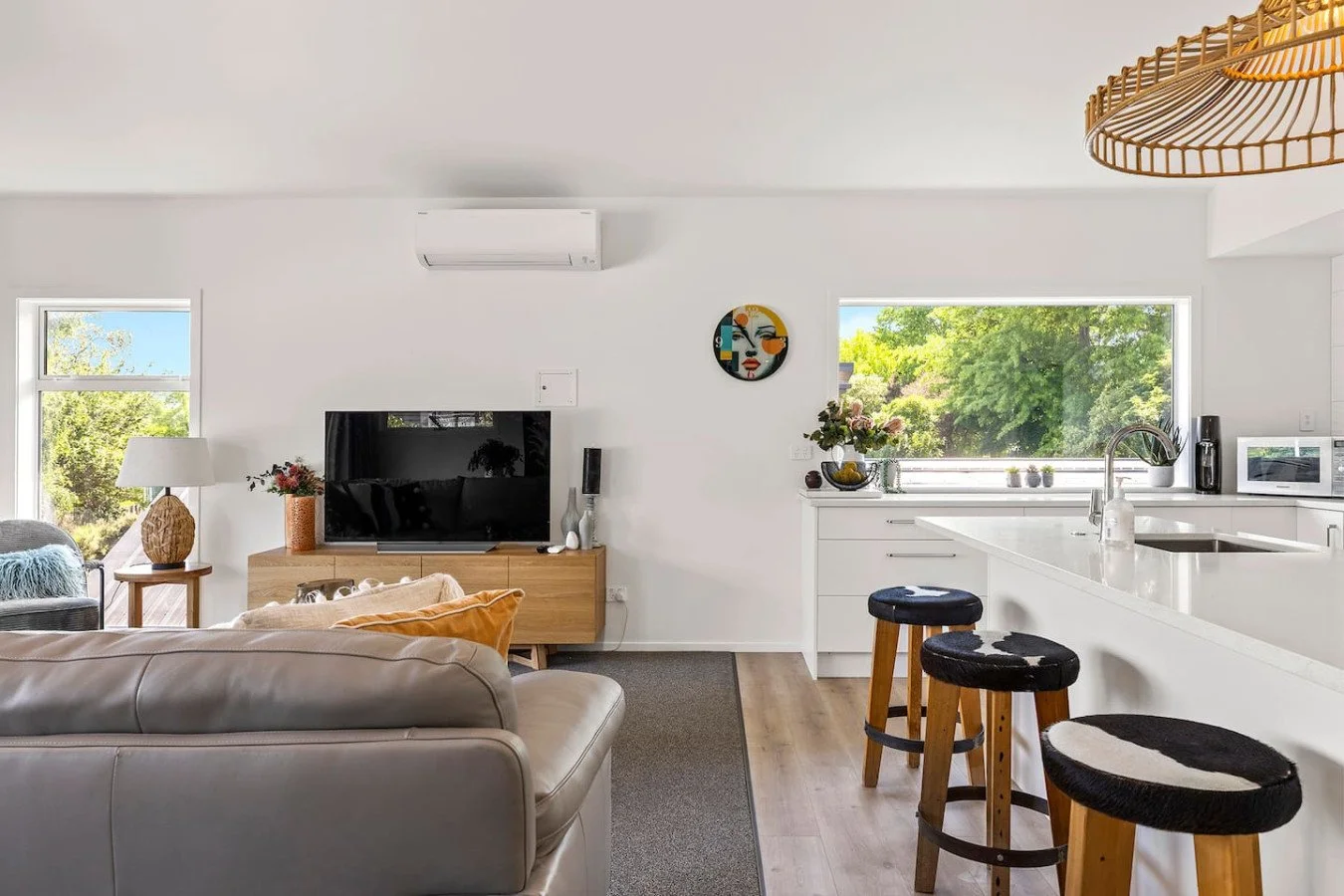 Modern living room and kitchen with white walls, a gray sofa, a flat-screen TV, a wooden TV stand, a lamp, a decorative vase, bar stools, countertops, a window with a view of trees, and minimal decor.