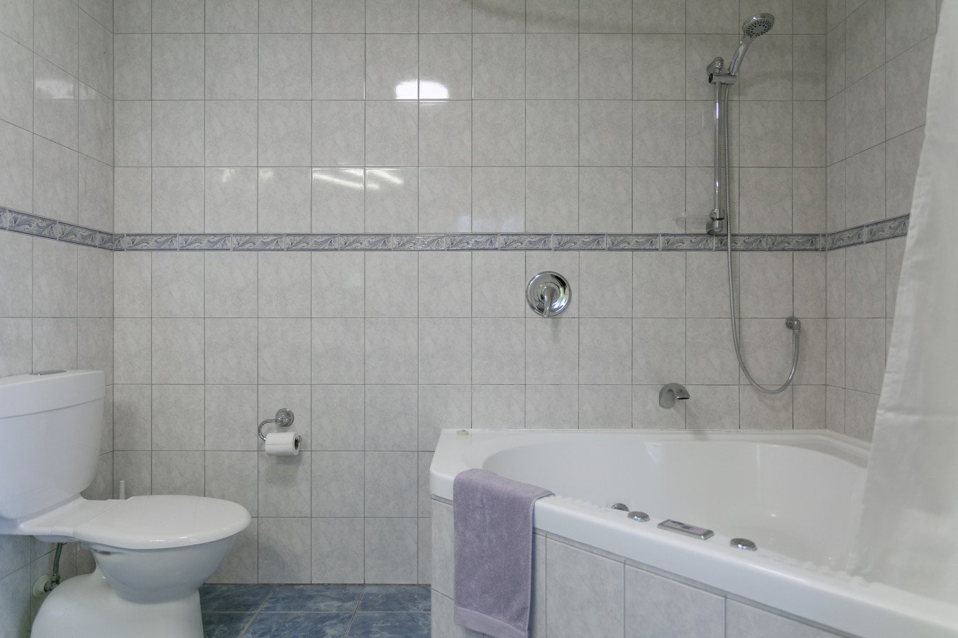 Bathroom with a toilet on the left, a bathtub with a towel hanging over the edge, a shower head, and tiled walls and floor.
