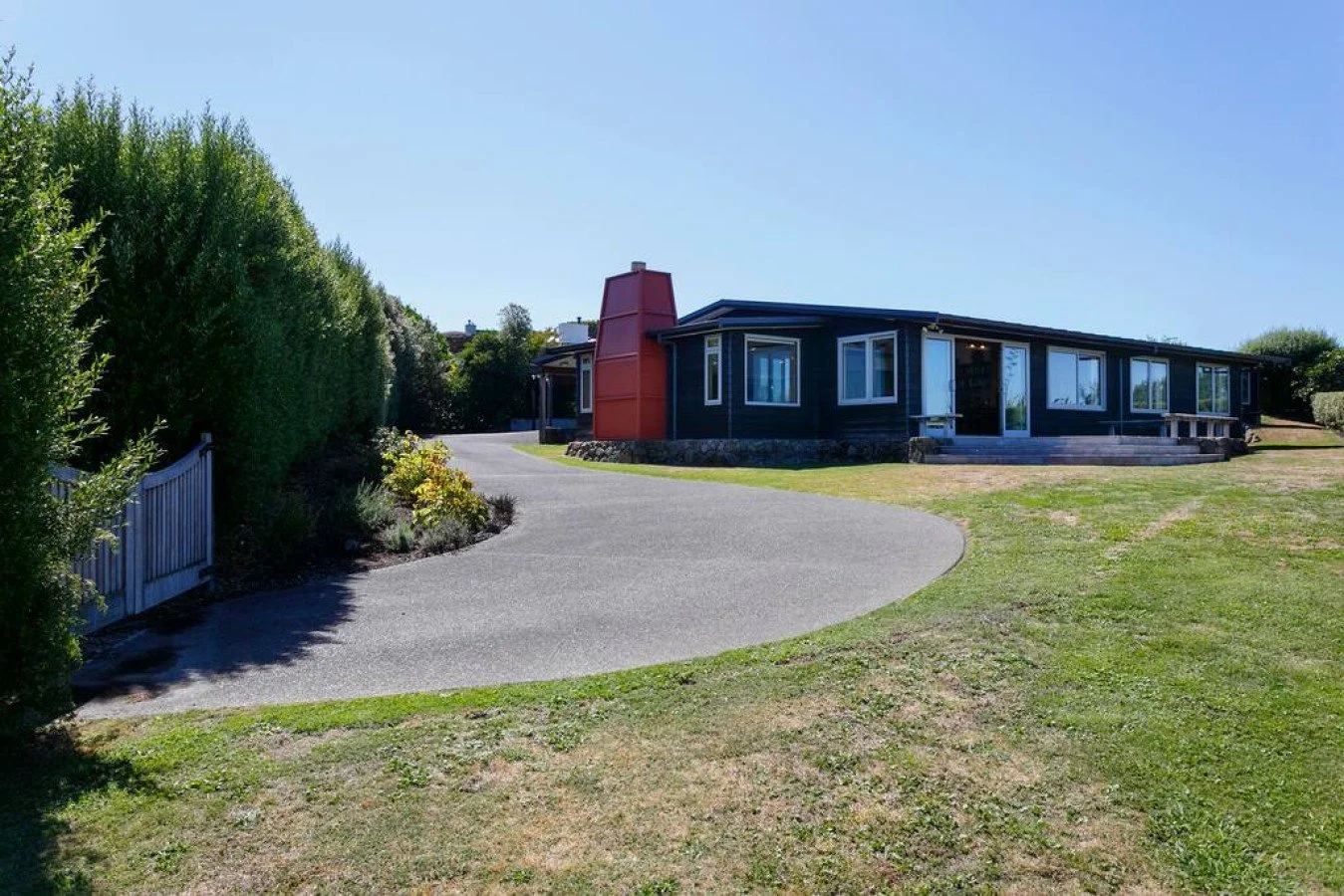 A modern black house with large windows and a red chimney, situated on a grassy landscape with a winding driveway and a white fence on the left.
