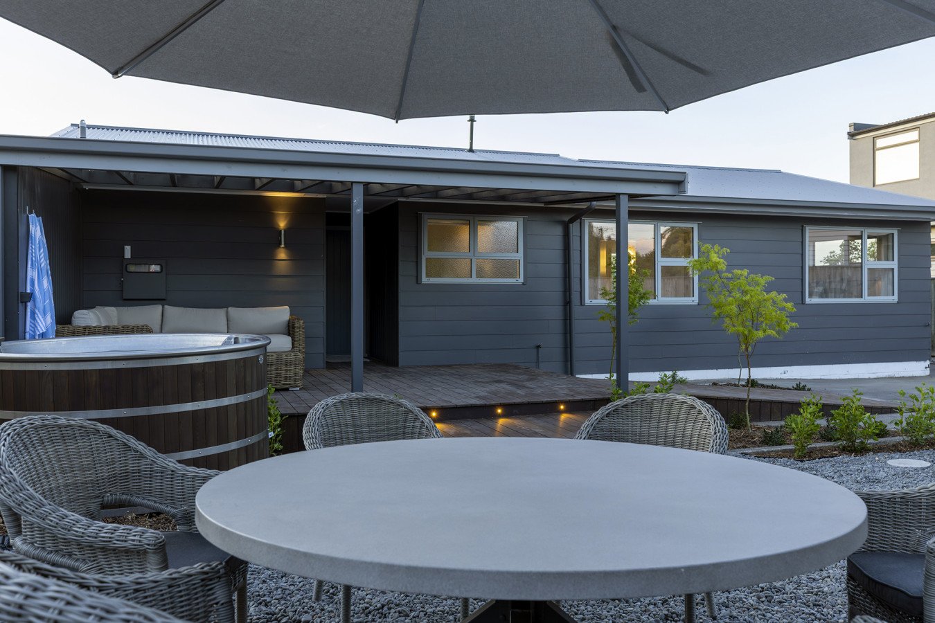 An outdoor patio area featuring a round table with wicker chairs, a hot tub, a large umbrella, and a house with blue siding, windows, and a small tree in front.