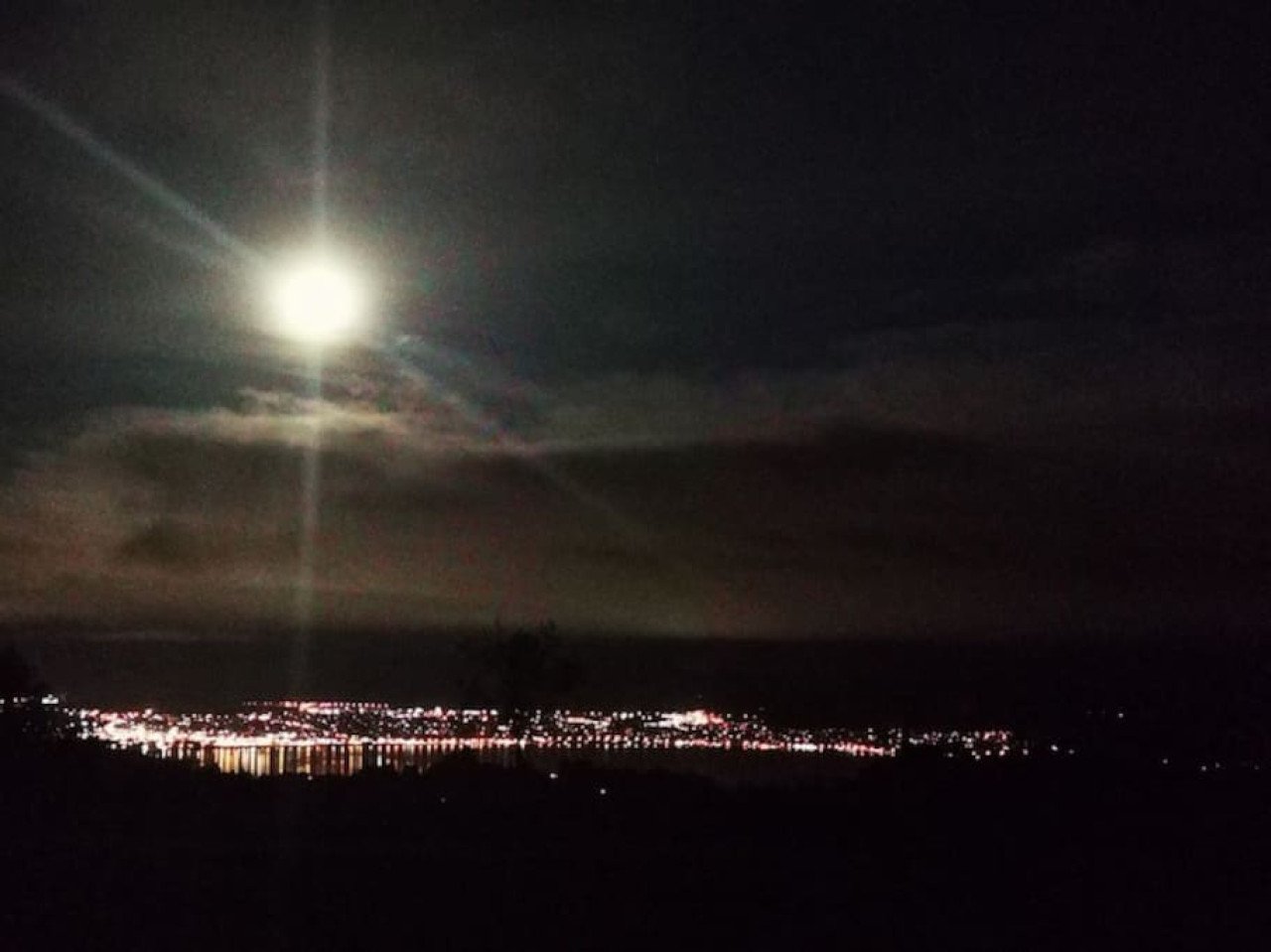 Nighttime scene with a bright full moon, some clouds, and a cityscape with scattered lights along a water body in the distance.