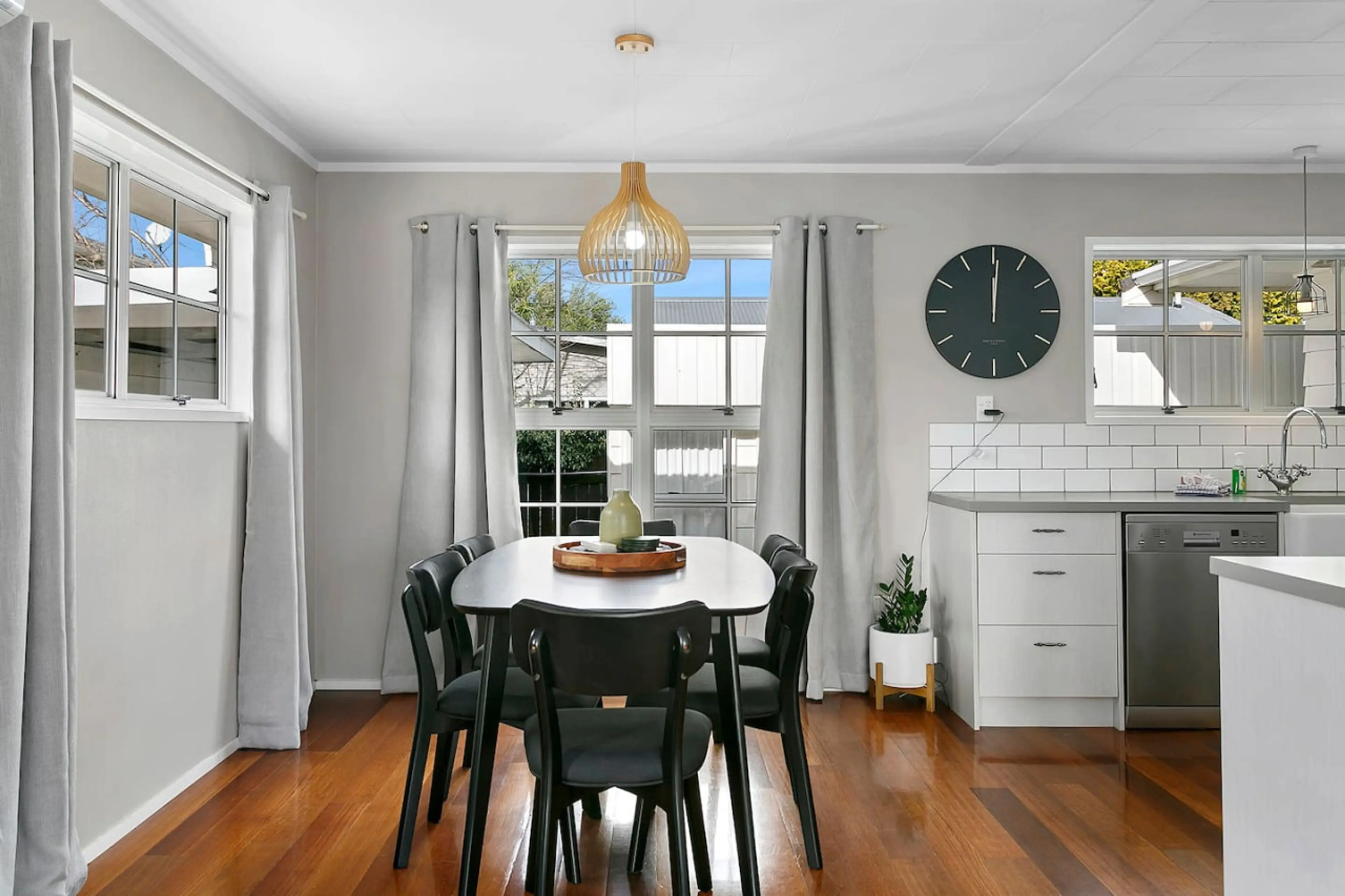 Dining area with a white table, black chairs, and a wooden tray with a pitcher and glasses. Large windows with white curtains, a wooden pendant light, and a black clock on the wall. Part of the kitchen with white cabinets, a plant, and a dishwasher.