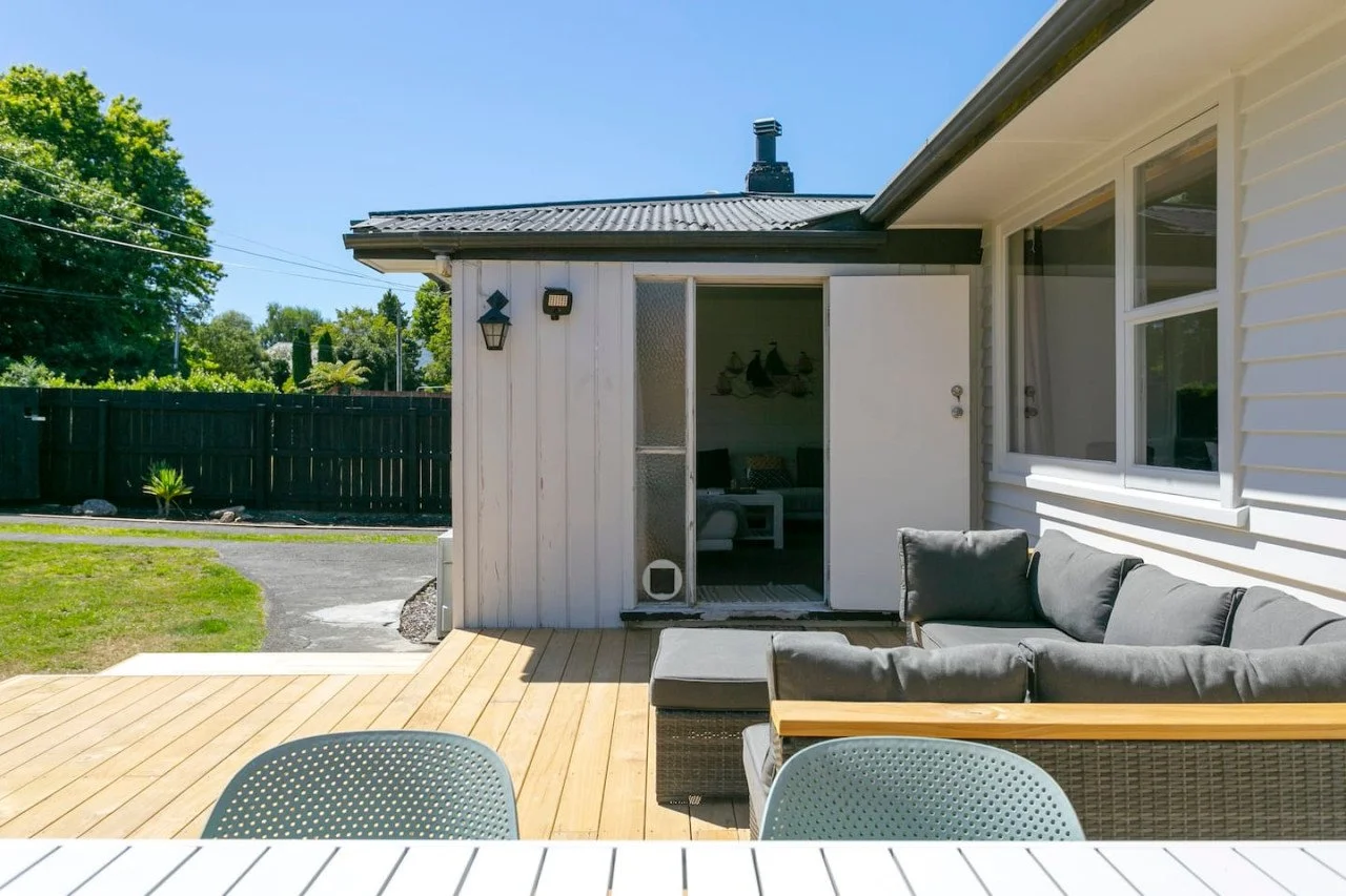 View of a backyard patio with outdoor sofa, white wooden deck, and exterior house wall with windows and sliding door, under clear blue sky.