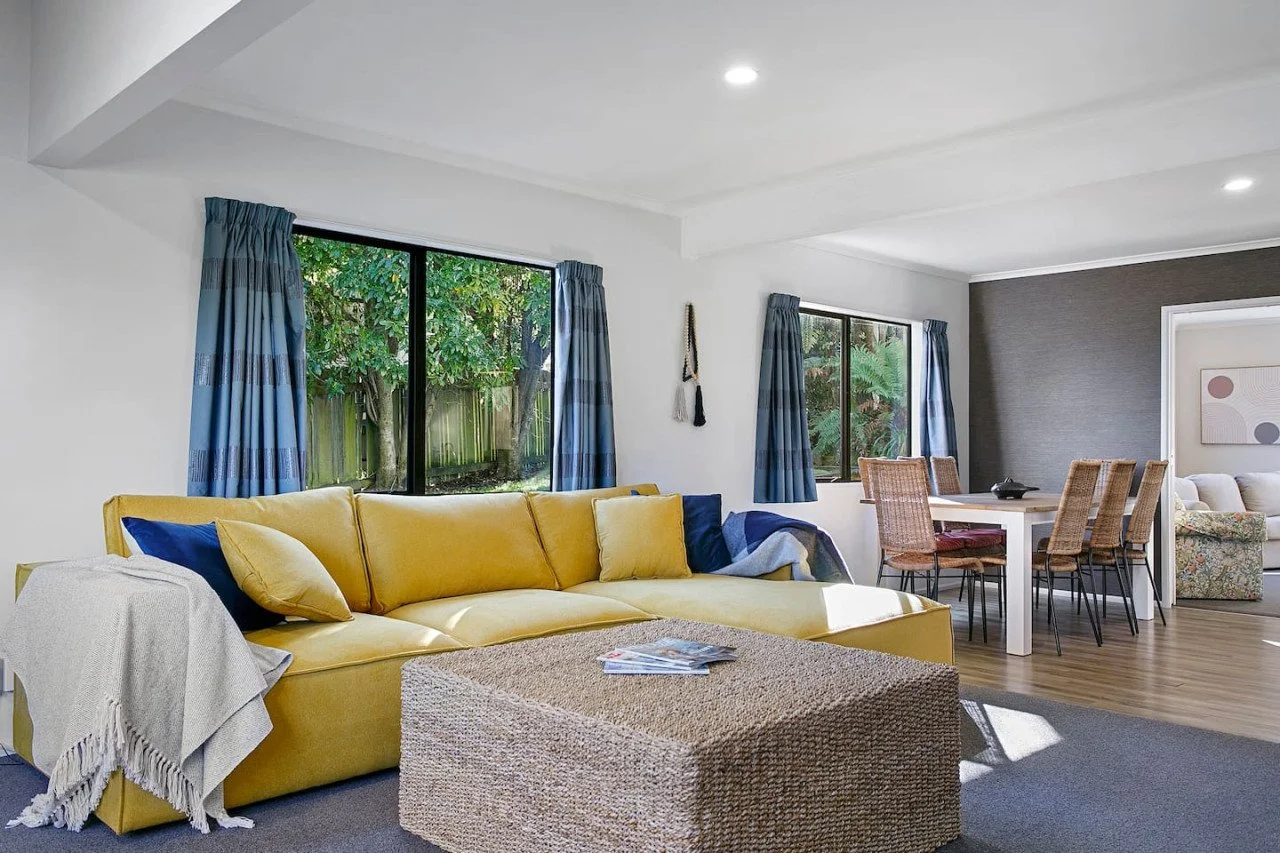 Bright living room with large yellow sofa, blue curtains, and a dining area with six chairs, next to windows showing greenery outside.