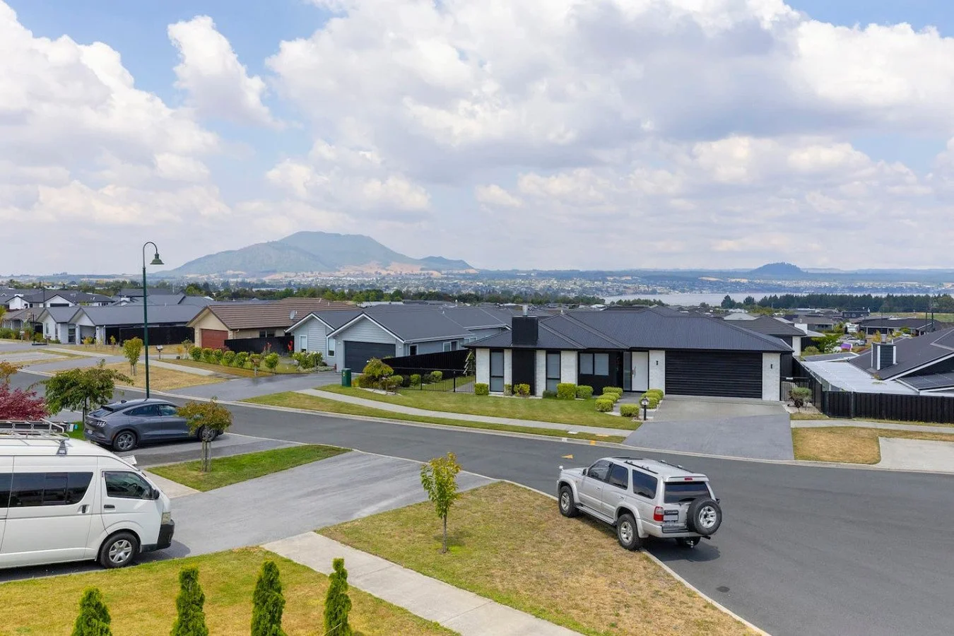 A suburban neighborhood with modern houses, parked cars, trees, a streetlamp, and a scenic background including mountains and water under a partly cloudy sky.