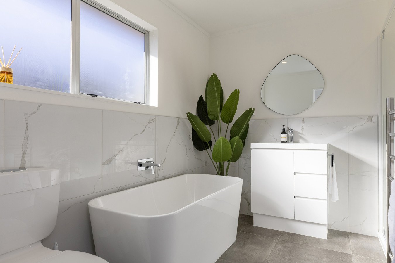 Modern bathroom with a white bathtub, a white vanity with a mirror, a potted plant, and a window allowing natural light.