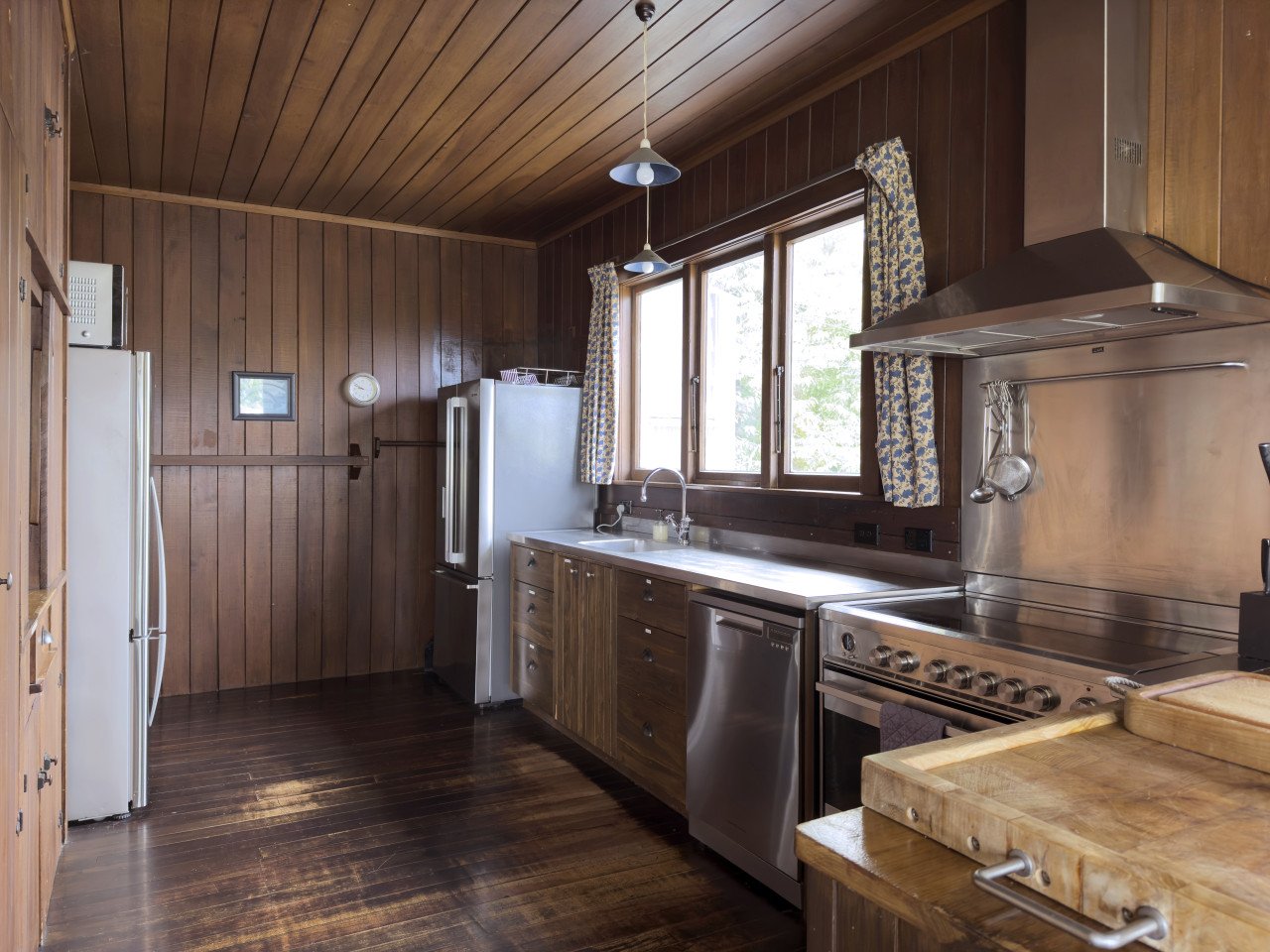 Wood-paneled kitchen with stainless steel appliances, wooden cabinetry, a window with patterned curtains, and a wooden countertop.