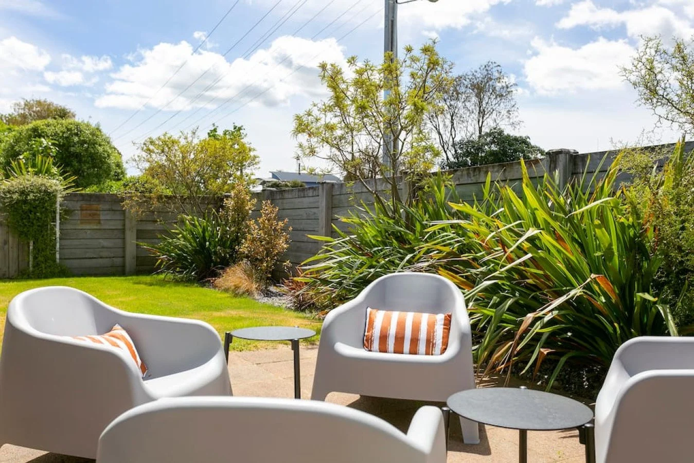 Outdoor patio seating with white chairs, small black tables, striped pillows, green plants, a wooden fence, and a partly cloudy sky.