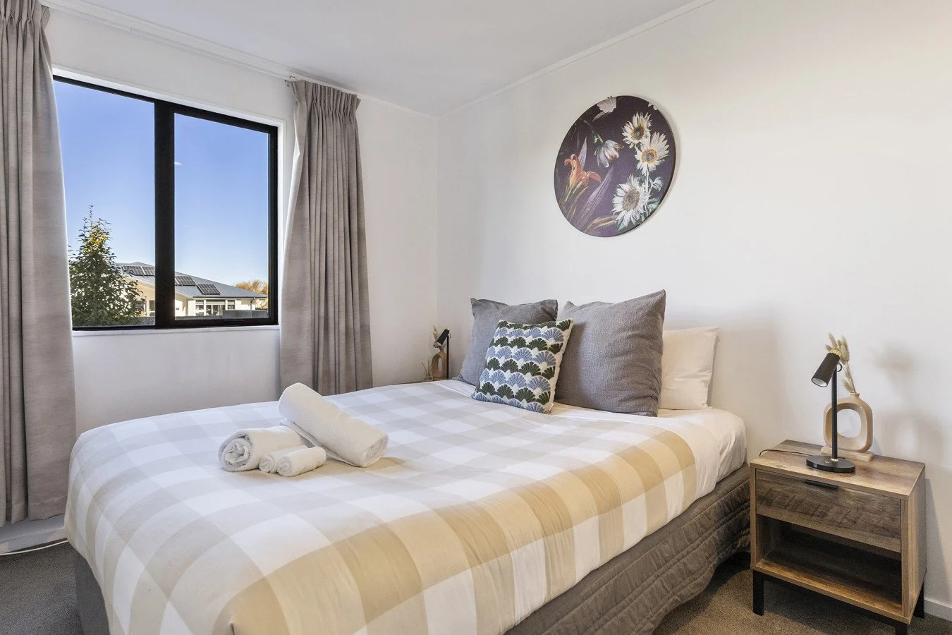 A tidy bedroom with a neatly made bed featuring a beige checkered duvet, three rolled white towels, and a decorative pillow. The room has a window with beige curtains, a nightstand with a black desk lamp, and a round floral wall art piece above the b