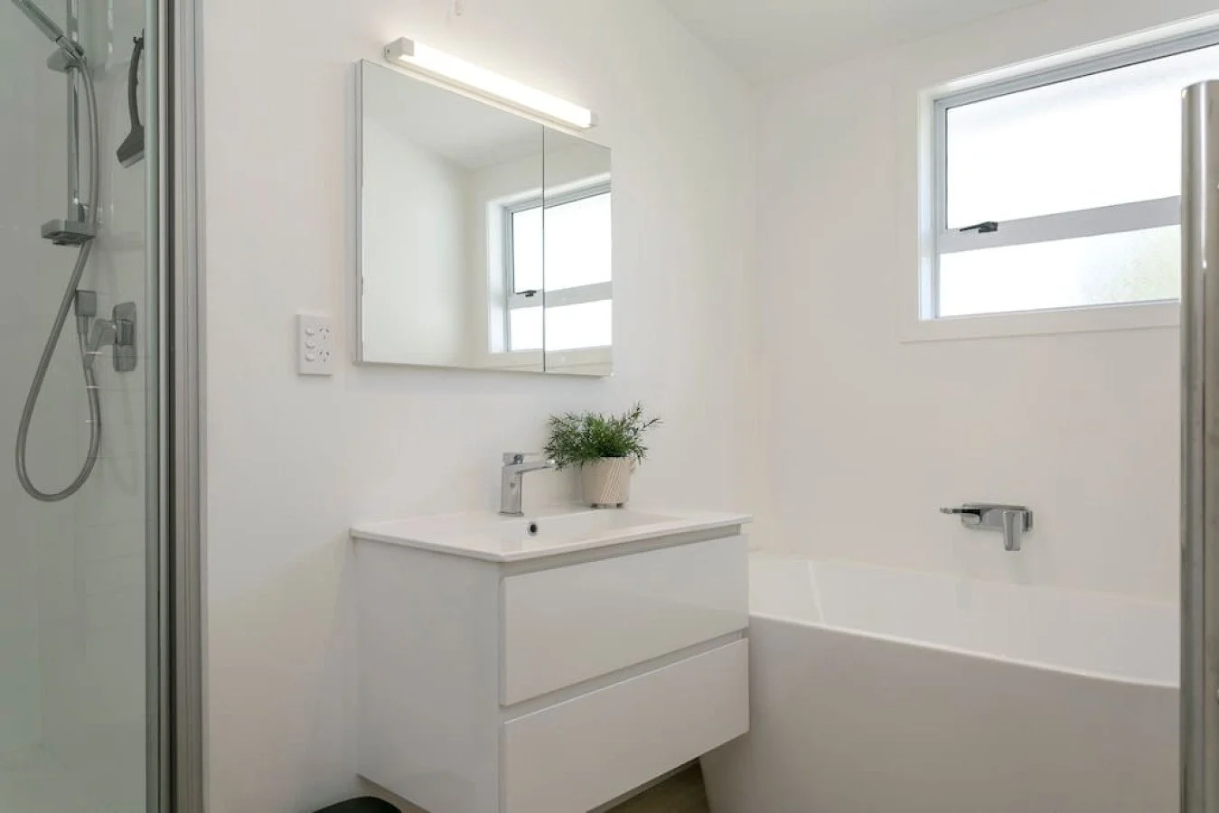 Bright white bathroom with a small vanity, mirror, potted plant, bathtub, window, and shower enclosure.