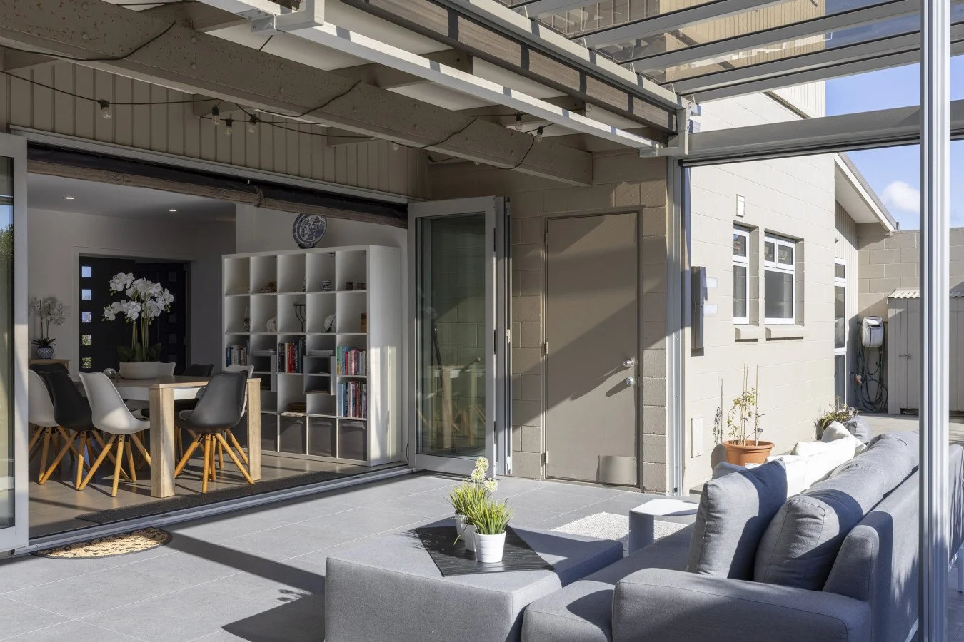 Outdoor patio with a gray couch, potted plants, and a view into a dining and living area inside a modern house.