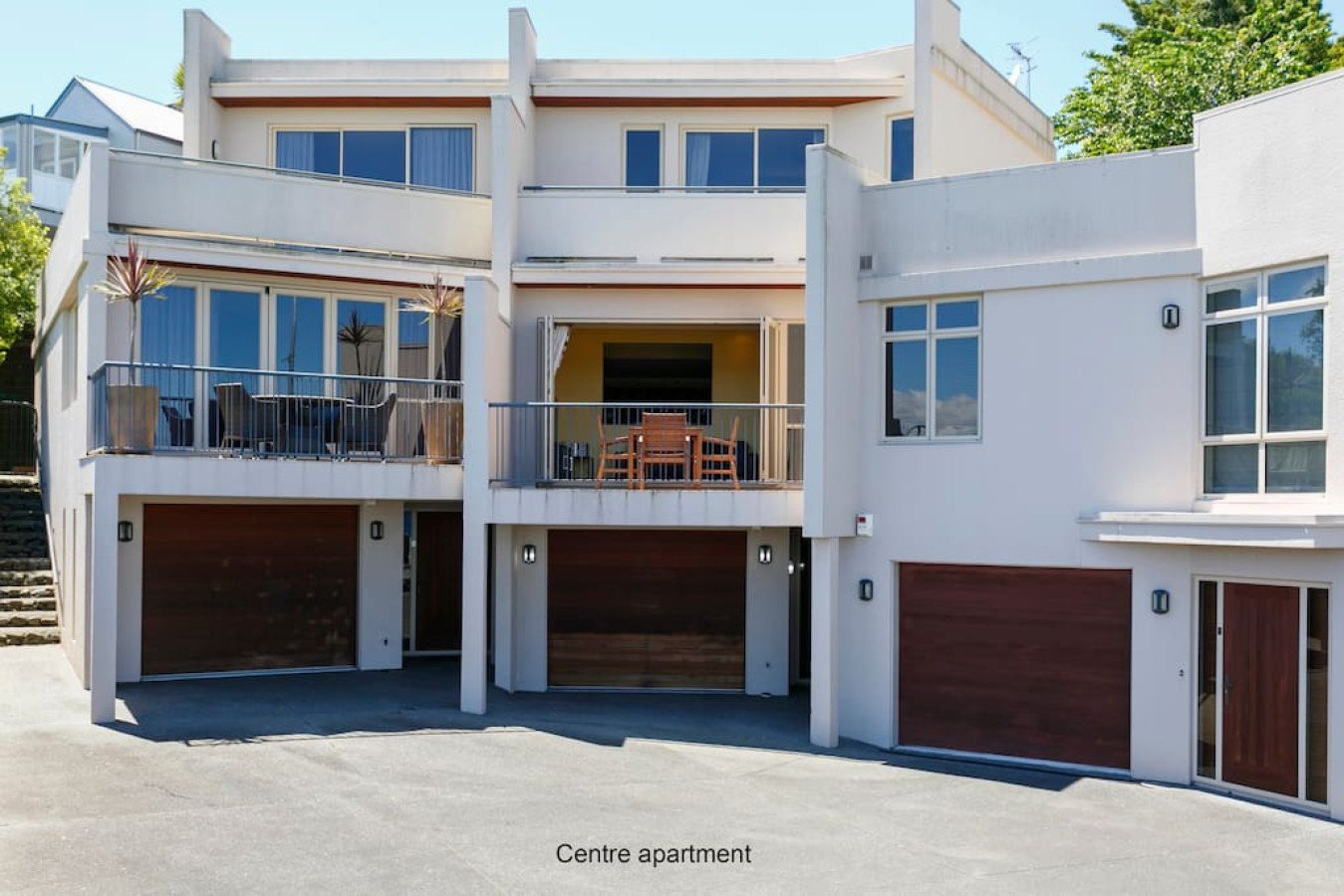 Modern white multi-story apartment building with balconies, large windows, and wooden garage doors. The building has outdoor patio furniture on the balconies and a small staircase to the left.