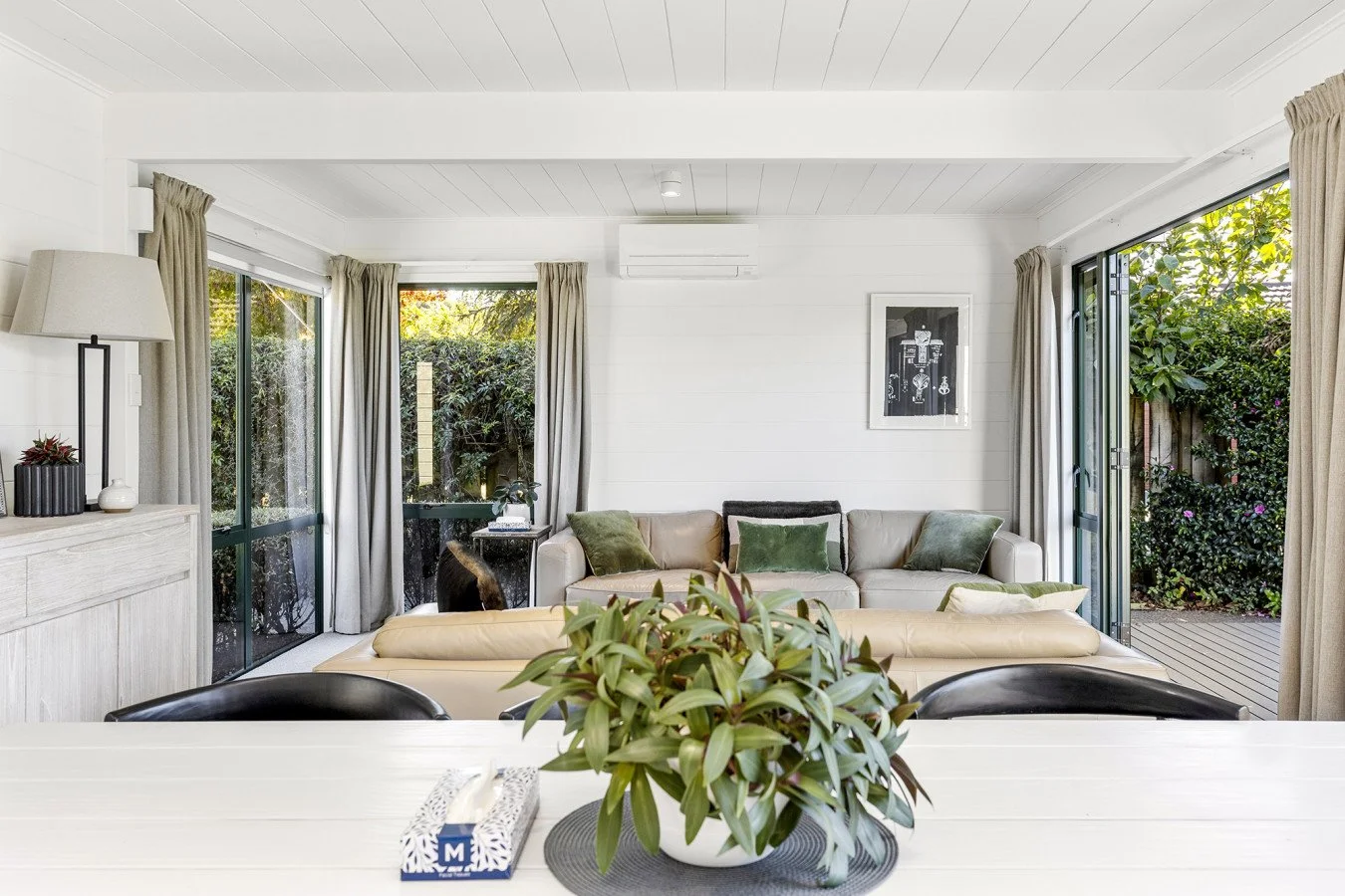 Living room with a beige sectional sofa, green cushions, and large windows showing greenery outside.