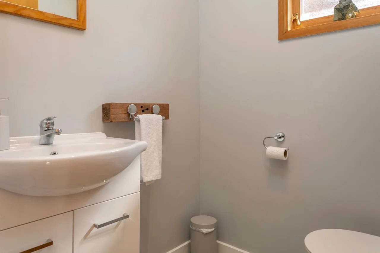 A small bathroom with a white sink, a white cabinet, a wooden towel rack with a towel, a small trash can, a toilet paper holder with a roll, and a window with wooden trim.