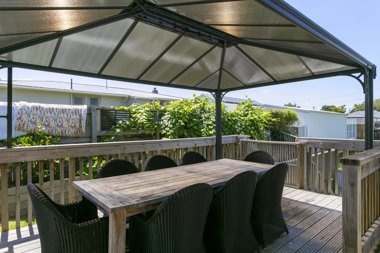Wooden deck with outdoor dining table and six wicker chairs, shaded by a metal patio cover, with lush greenery and a fence in the background.