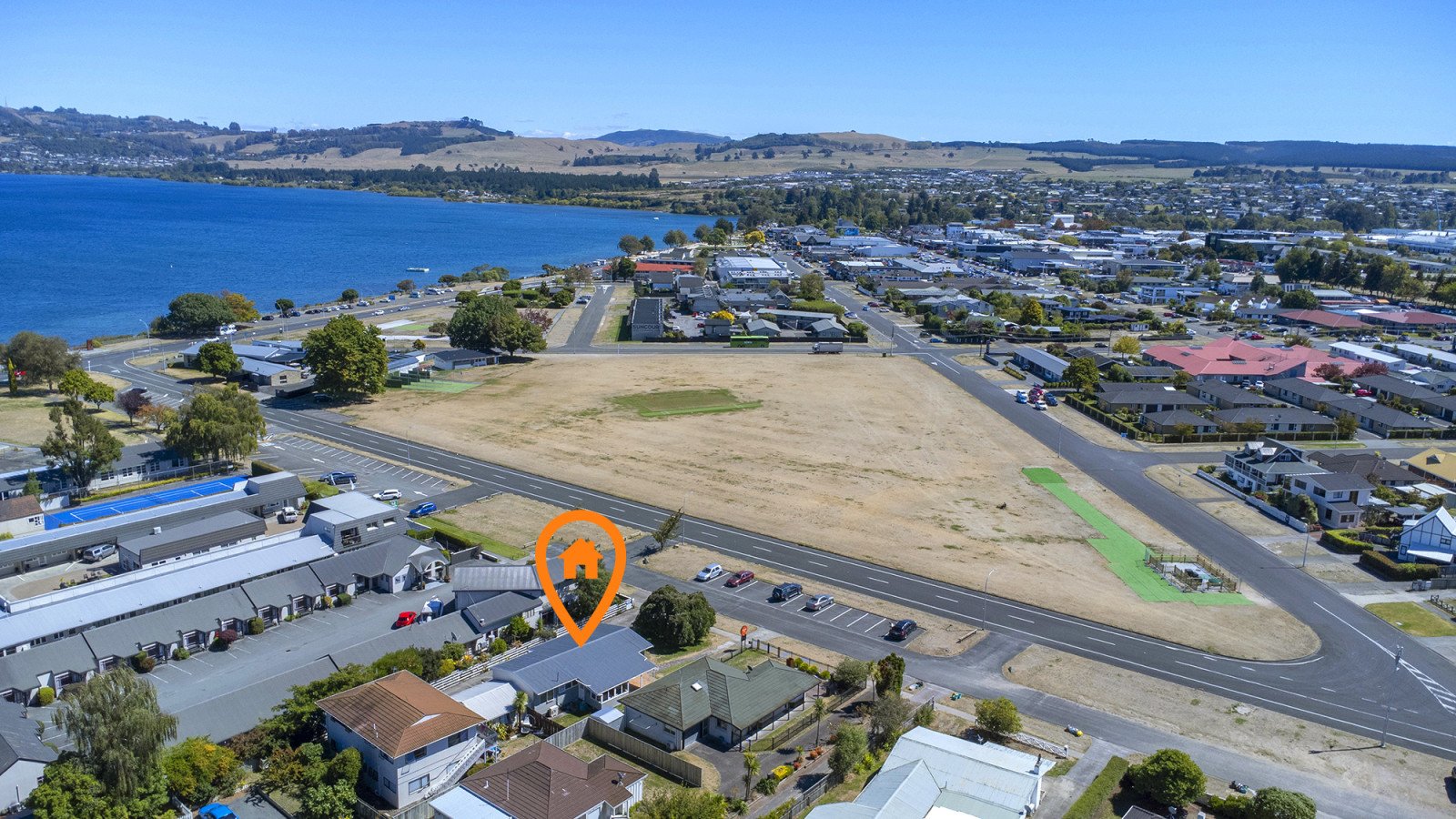 Aerial view of a residential neighborhood near a lake, with an empty field in the center, marked with an orange house icon.