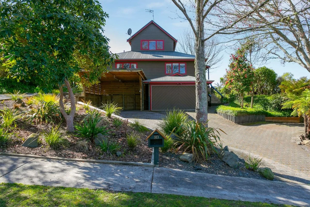 A multi-story house with grey siding and red window frames, surrounded by trees and landscaped garden, with a driveway leading to a garage.