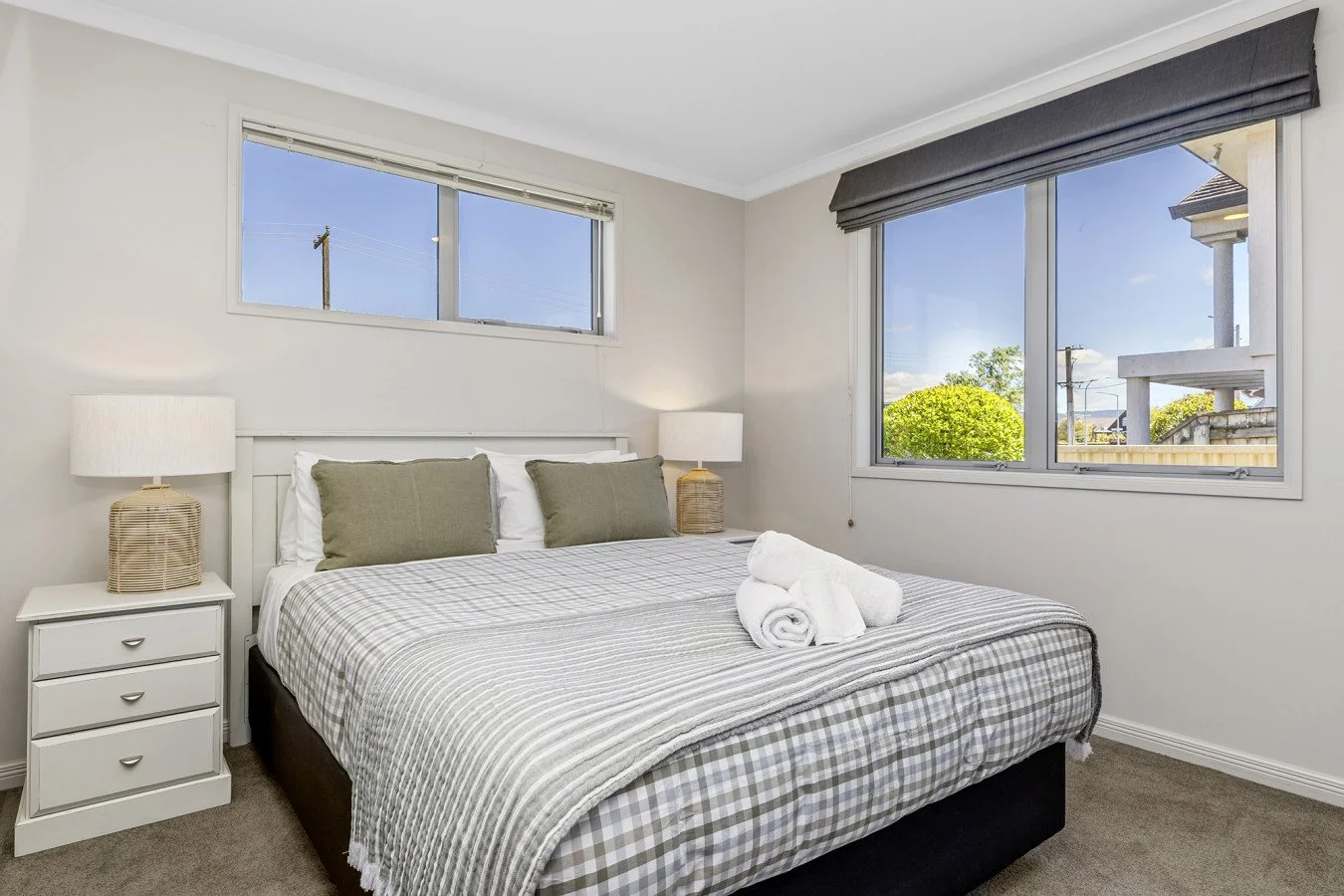 A bedroom with white walls, two large windows showing blue sky and greenery, a bed with white and gray checkered bedding, two green pillows, two white rolled towels on the bed, and two bedside tables with lamps.