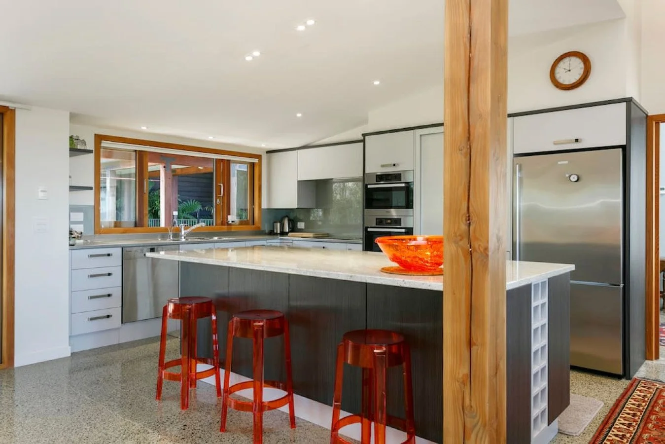 Modern kitchen with white cabinets, a large island with granite countertop, red bar stools, stainless steel refrigerator, wall oven, and a window overlooking a patio.