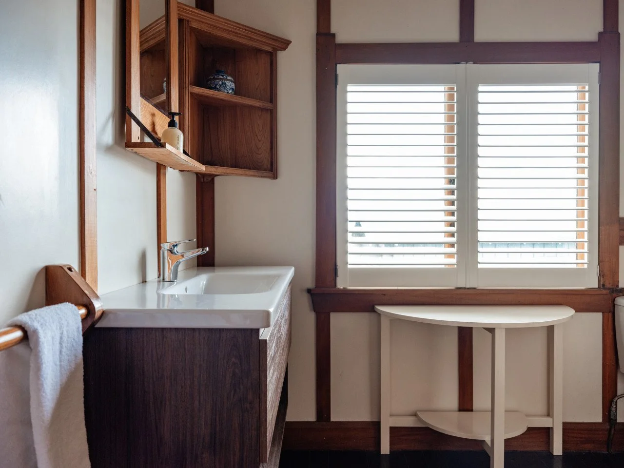 Bathroom with a white sink, wooden shelves, a window with white blinds, and a small white table.