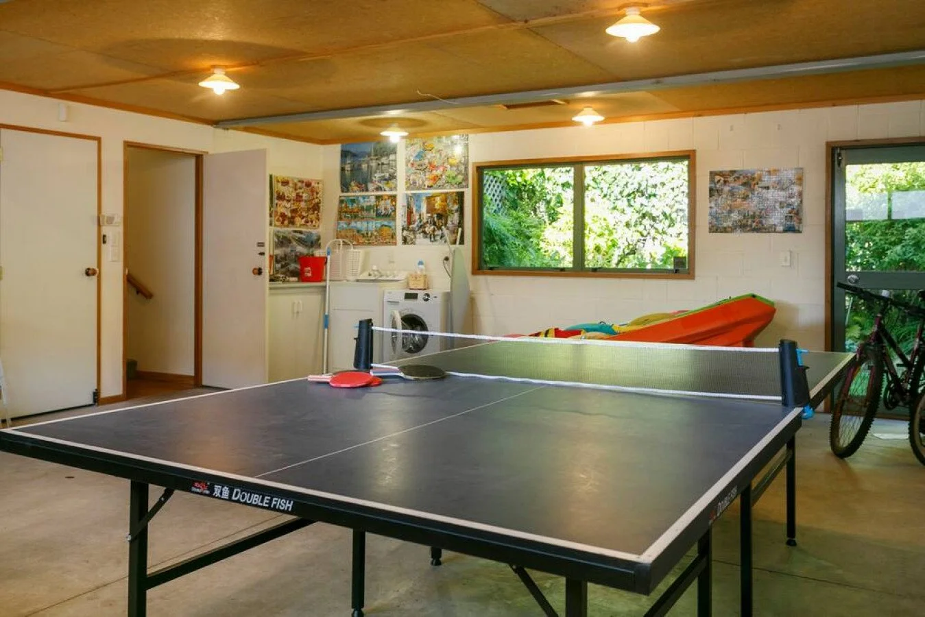 Indoor recreational room with a ping pong table, a bicycle, a canoe, a washing machine, and laundry supplies. The room has wooden ceiling panels, white walls with colorful artwork, a large window showing green foliage outside, and a door leading to a