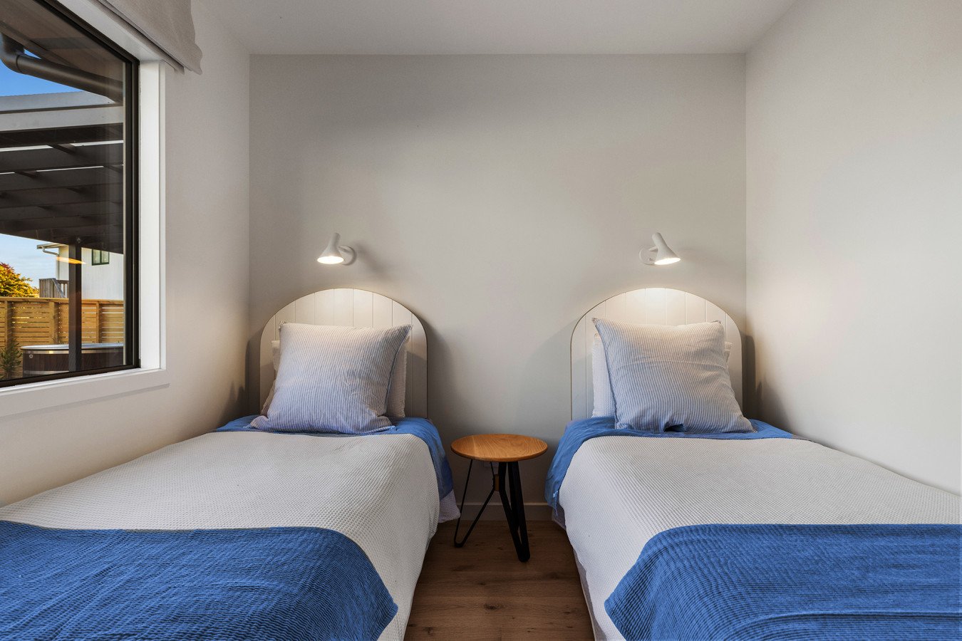 Room with two twin beds, white headboards, matching pillows and bedding, small wooden table between the beds, wall-mounted reading lights, window showing outside view.