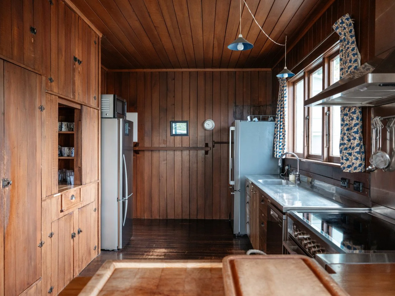 A rustic kitchen with wooden paneled walls and ceiling, featuring a refrigerator, microwave, stove, and a sink beneath three windows with patterned curtains, decorated with a small picture and a clock on the back wall.