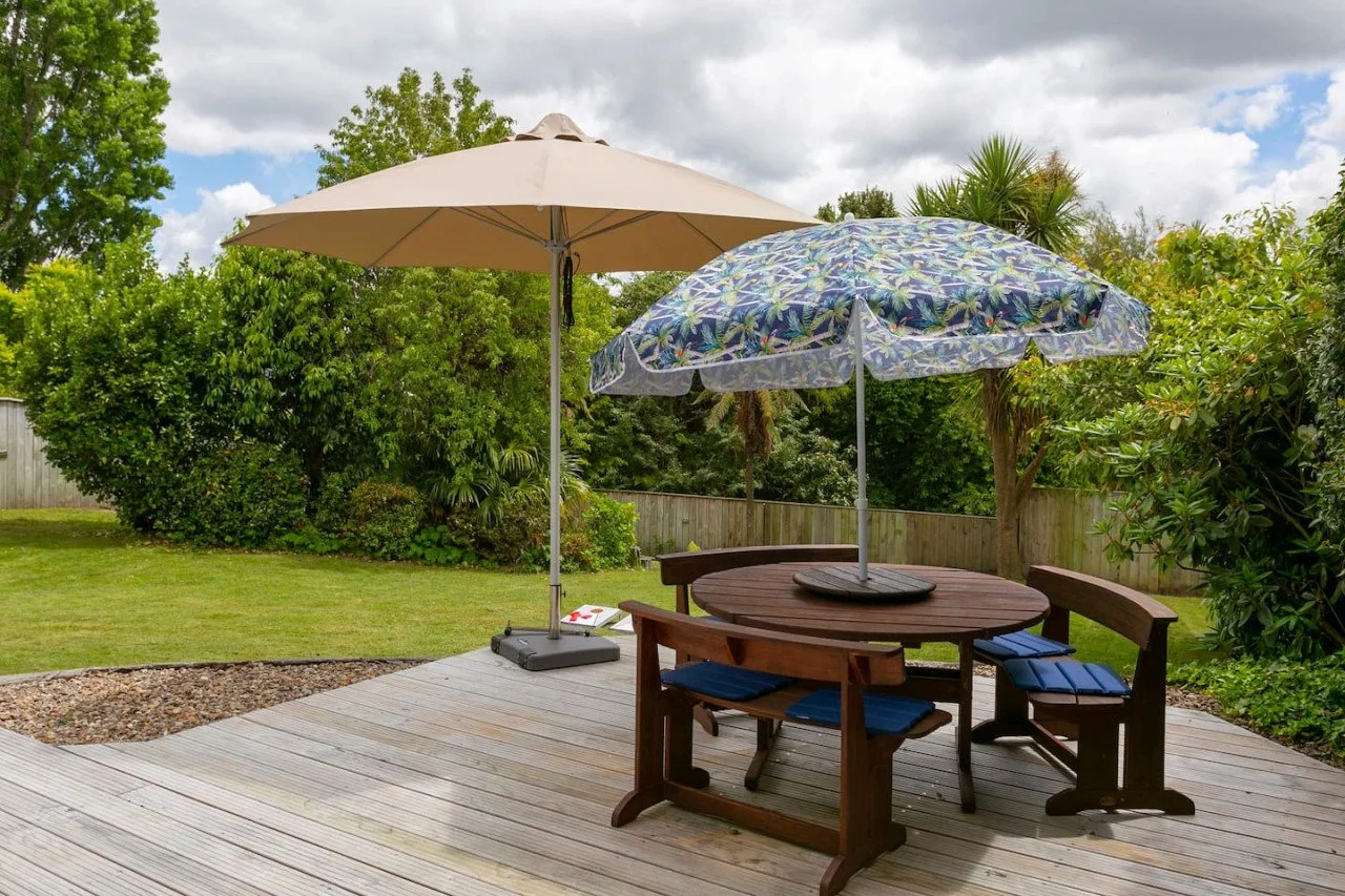 A backyard with a wooden deck, two patio umbrellas (one beige and one with a tropical floral pattern), a round wooden table, and four chairs beneath the umbrellas. Green bushes, trees, and a wooden fence surround the yard under a partly cloudy sky.