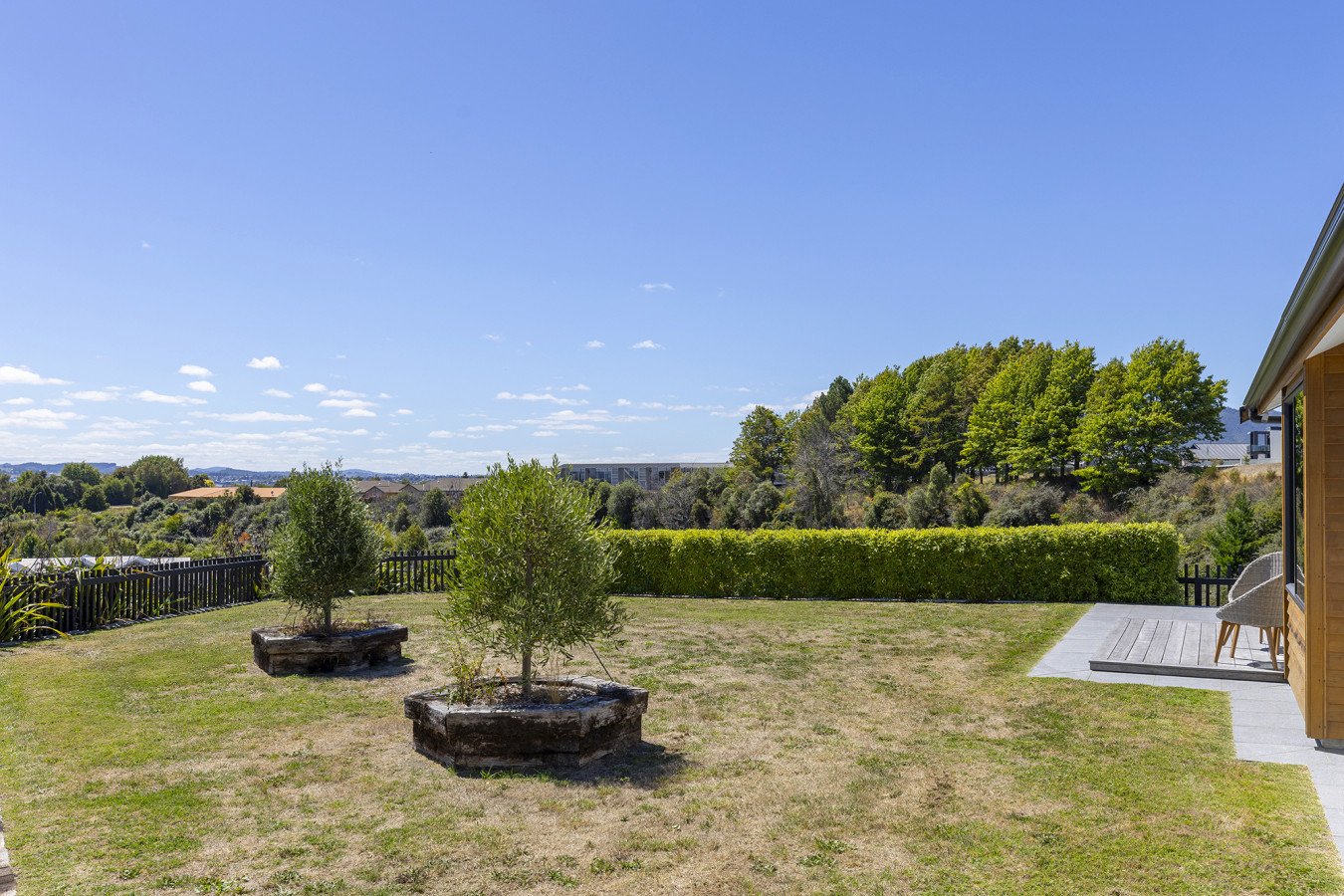 A backyard with a wooden deck, two chairs, a trimmed hedge, two small trees in planter boxes, a grassy lawn, and a distant view of trees and blue sky with some clouds.