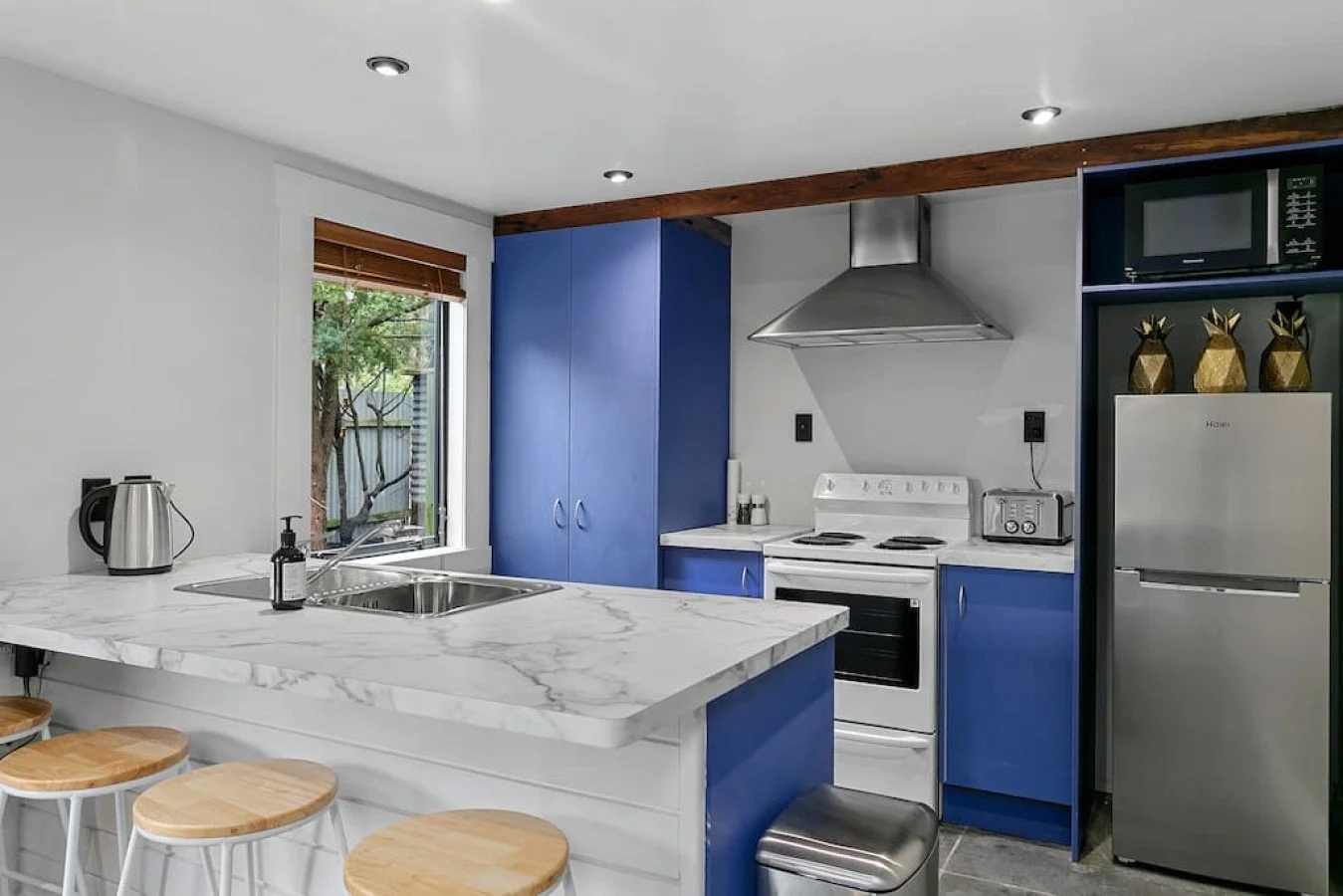 Modern kitchen with white and blue cabinetry, stainless steel appliances, and a marble countertop island with three wooden stools.