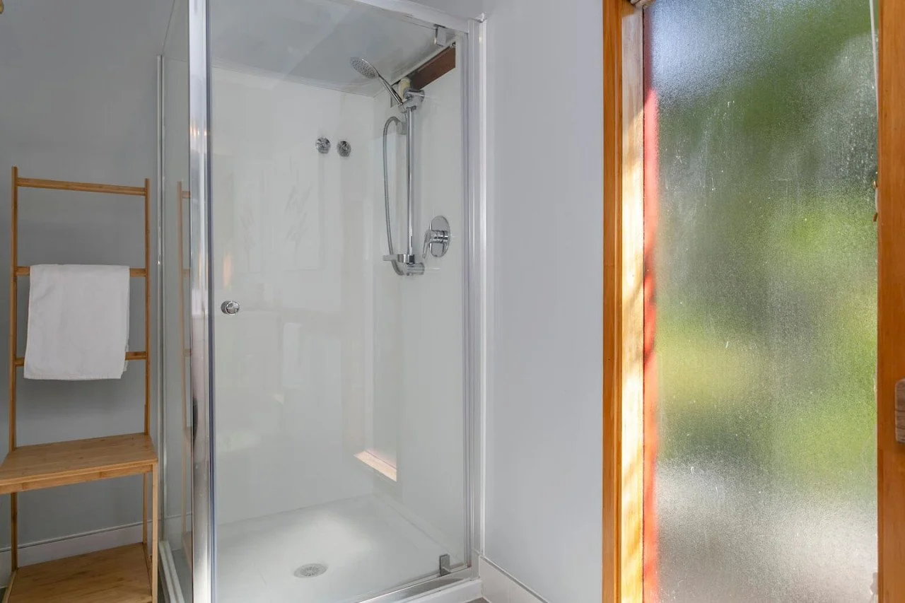Empty shower stall with a handheld showerhead and a glass door, next to a wooden towel rack with a white towel, and a frosted window with a wooden frame on the right.