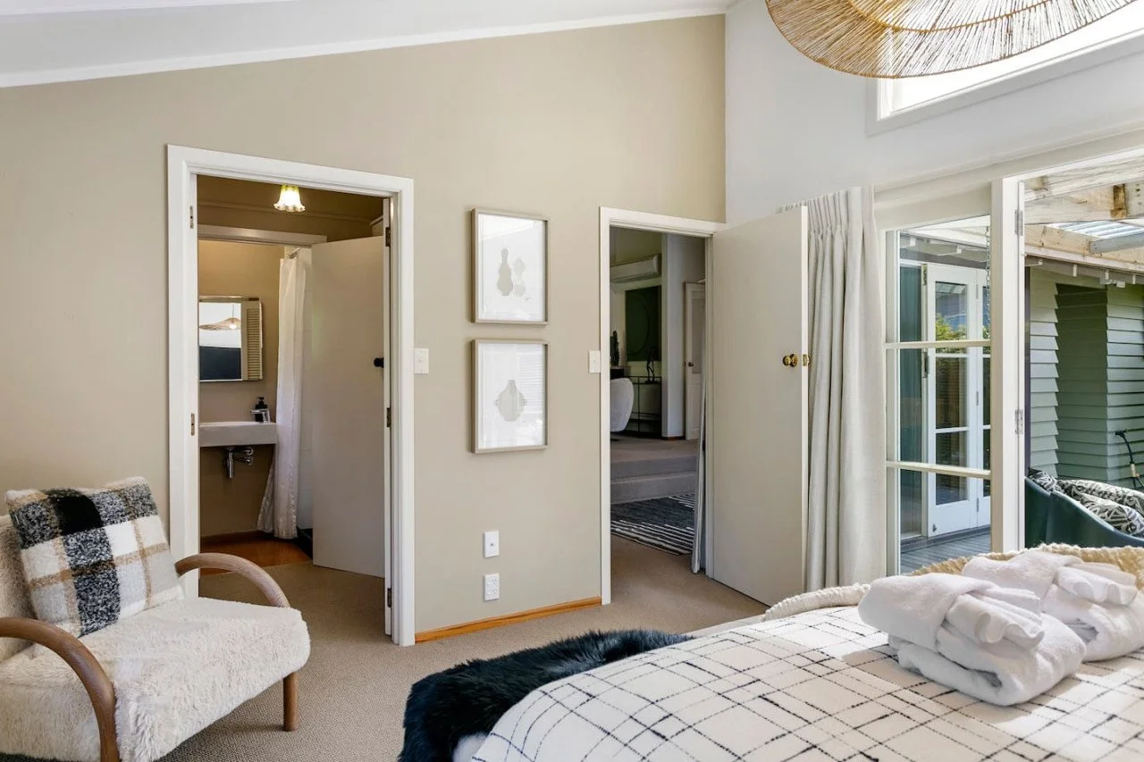 A cozy bedroom with a large bed covered with a checked blanket, white robes folded at the foot, and black fur throw. To the left, a beige armchair with a plaid pillow. Behind the bed, a dual glass door leads to a small balcony with outdoor furniture.