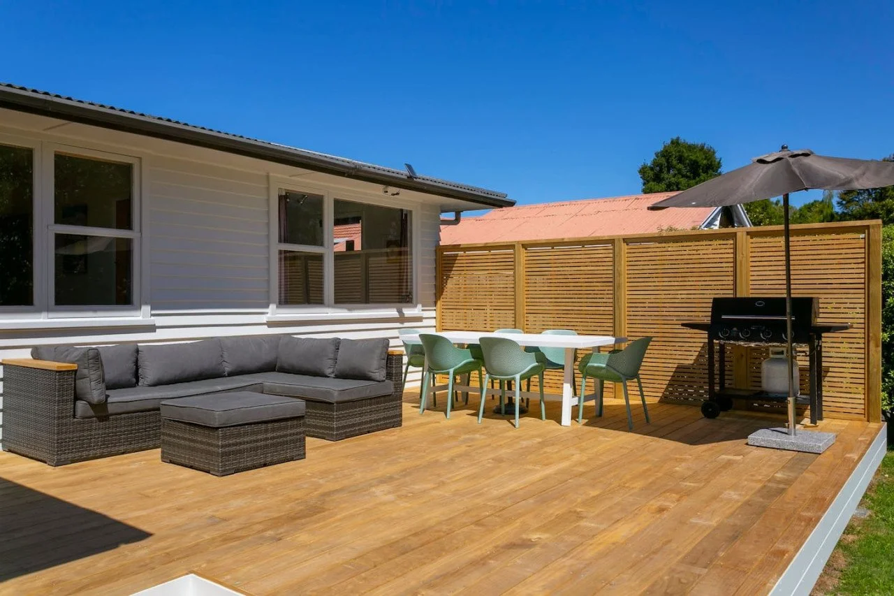 Wooden outdoor patio with gray sectional sofa, green chairs, white table, black grill with umbrella, and wooden privacy fence under a clear blue sky.