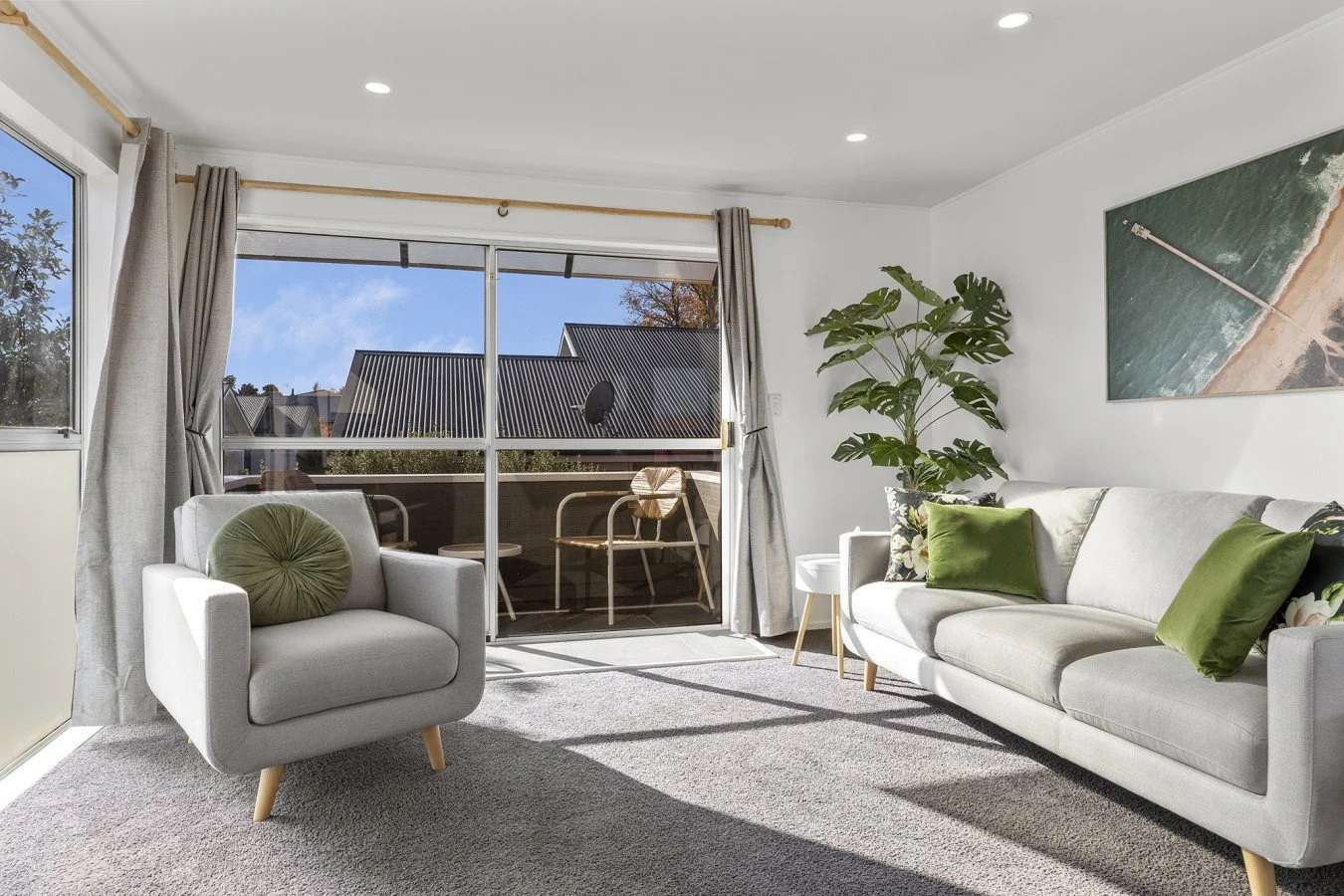 Living room with white sofa and armchair, green cushions, large window with curtains, potted plant, wall art, and a balcony with outdoor furniture.