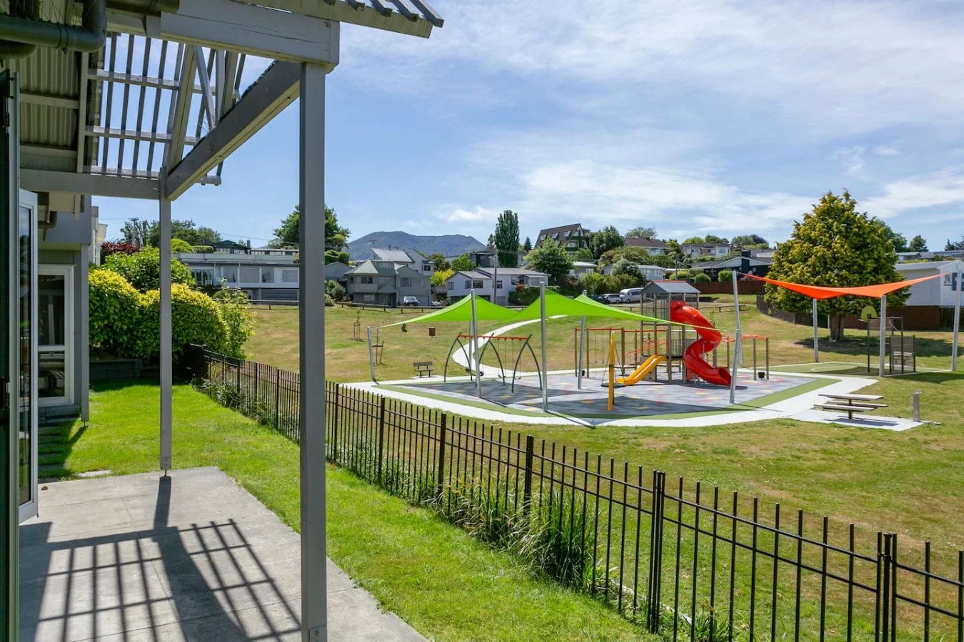 View of a playground with a red slide, swings, and shaded area, surrounded by a small fence, with houses and mountains in the background on a sunny day.