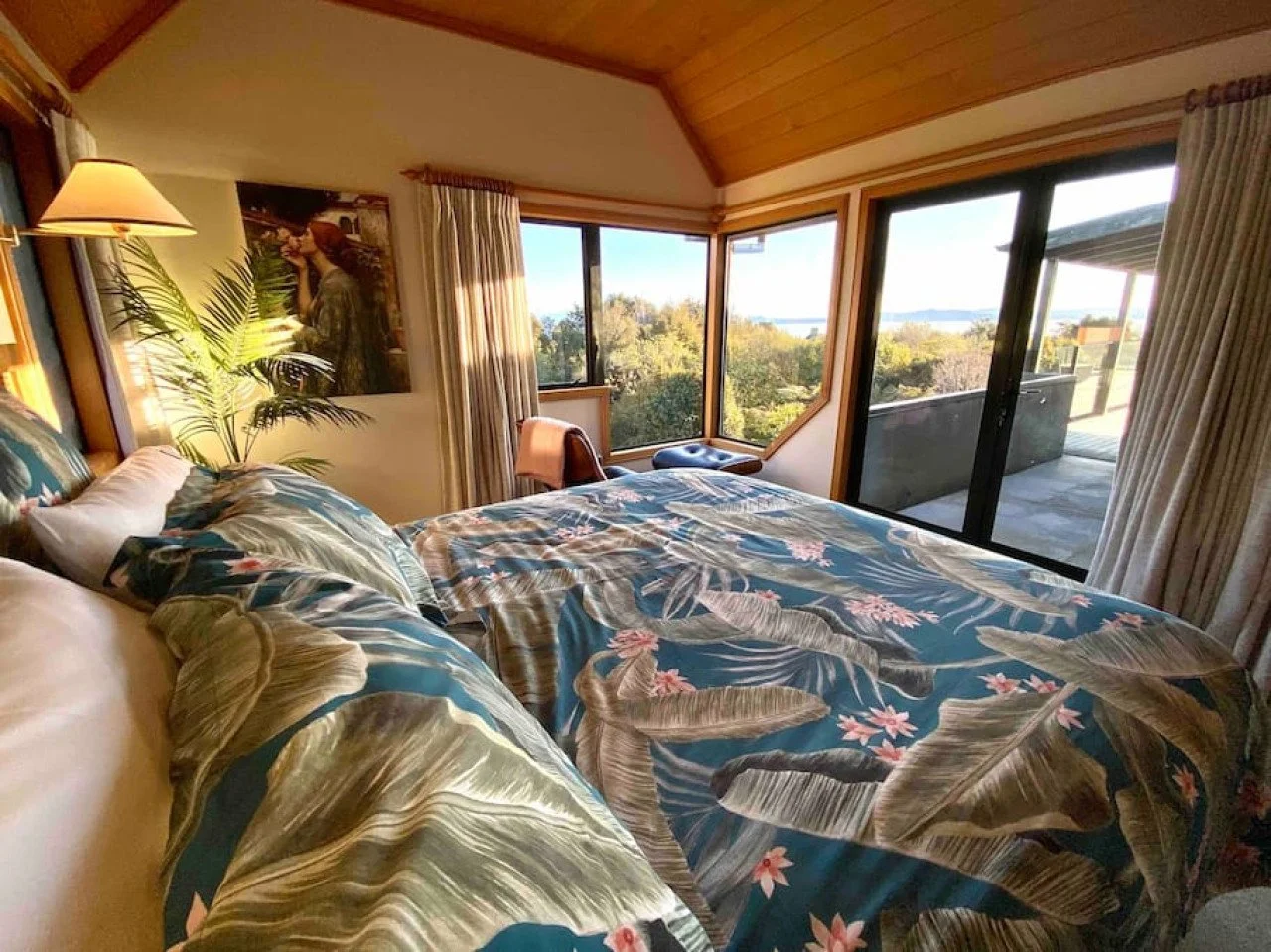 Cozy bedroom with large windows, tropical-themed bedding, a potted plant, and a balcony overlooking a scenic landscape.