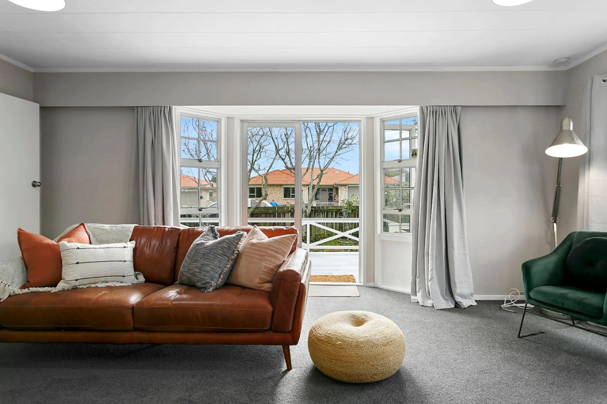 Living room with a brown leather sofa, decorative pillows, green armchair, tall floor lamp, and open doors leading to a porch with trees and houses outside.