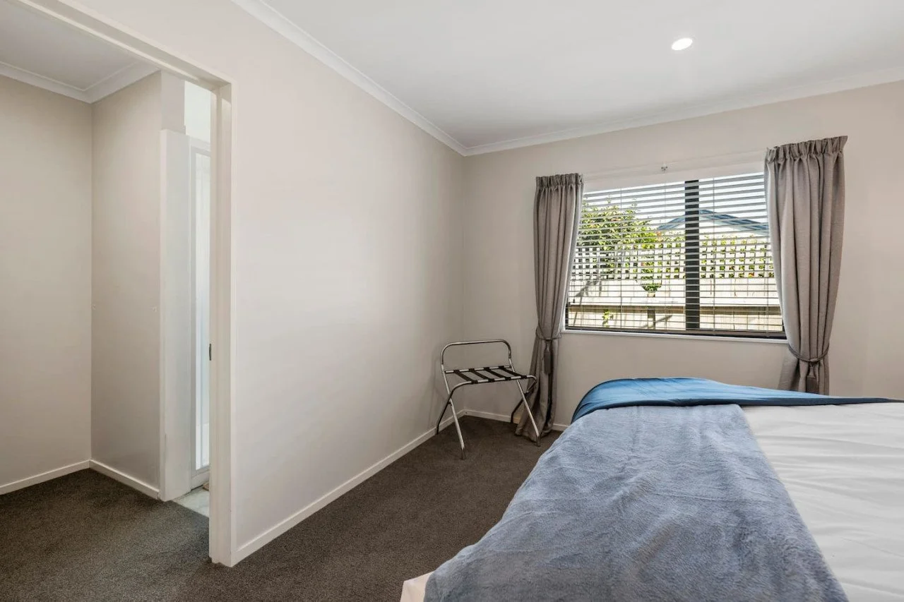 A simple bedroom with a bed partially shown, window with blinds and curtains, a luggage rack in the corner, and a door leading outside. The room has neutral-colored walls and carpeted floor.
