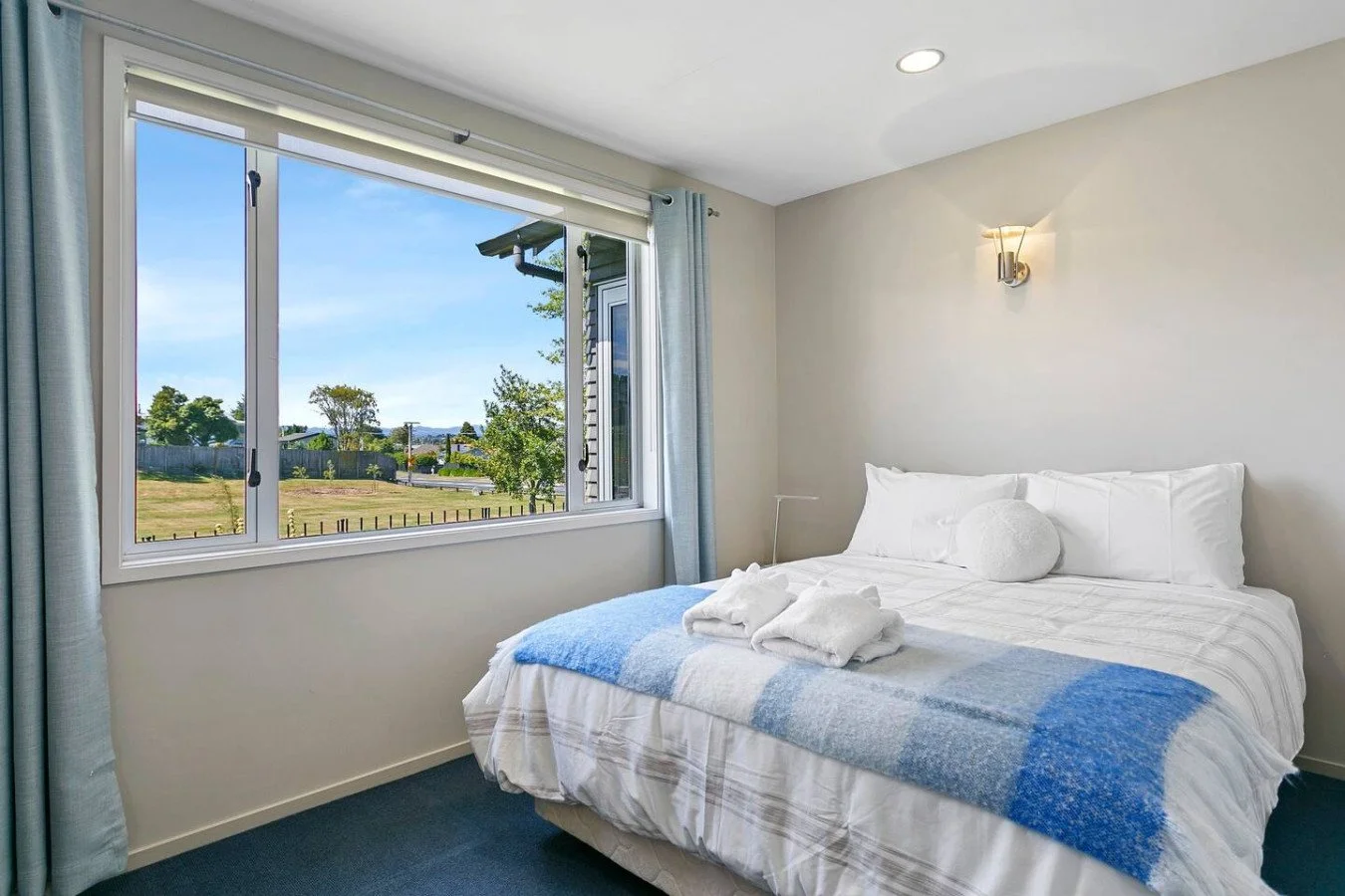 Bedroom with large window showing a view of trees and a grassy yard, white bed with blue and white blanket, white pillows, and folded towels on the bed, soft lighting from wall sconce.