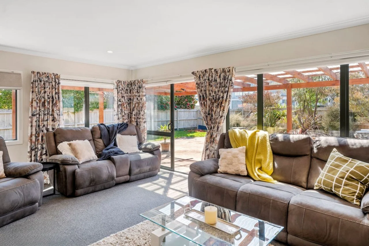 Living room with large windows, brown leather couches, colorful curtains, and a glass coffee table with candles.